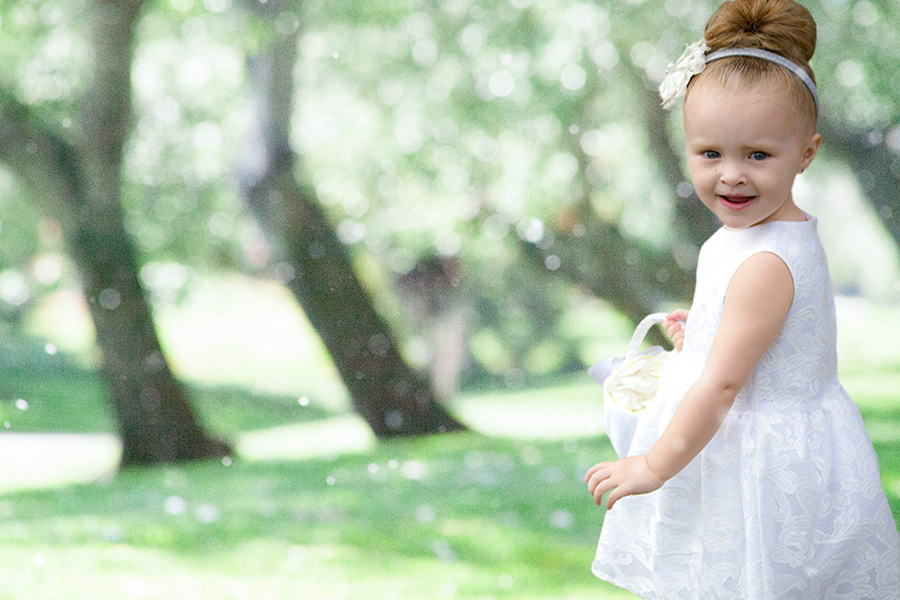 Lifestyle photography of toddler girl holding basket of flower pedals in front of tree wearing white dress