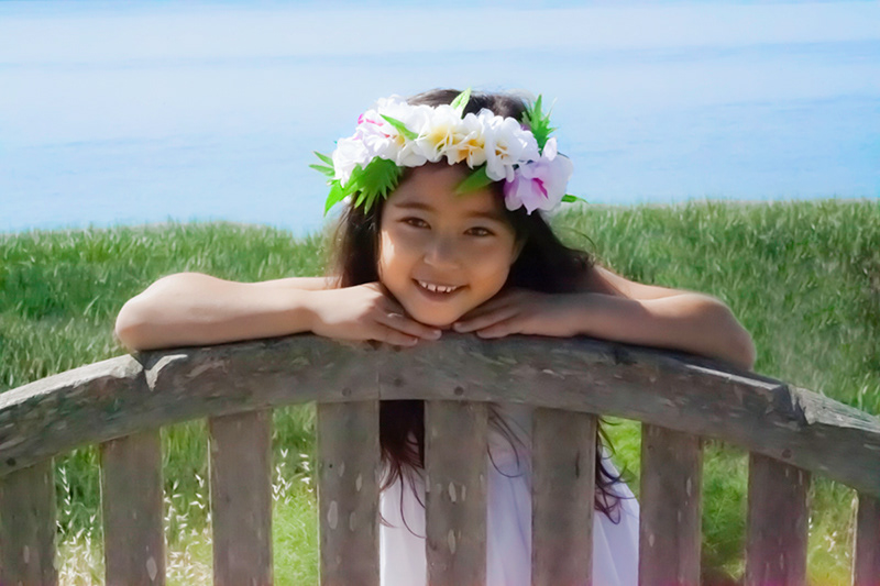 Photo of young girl wearing Hawaiian lei with hands on top of bench in front of blue waters