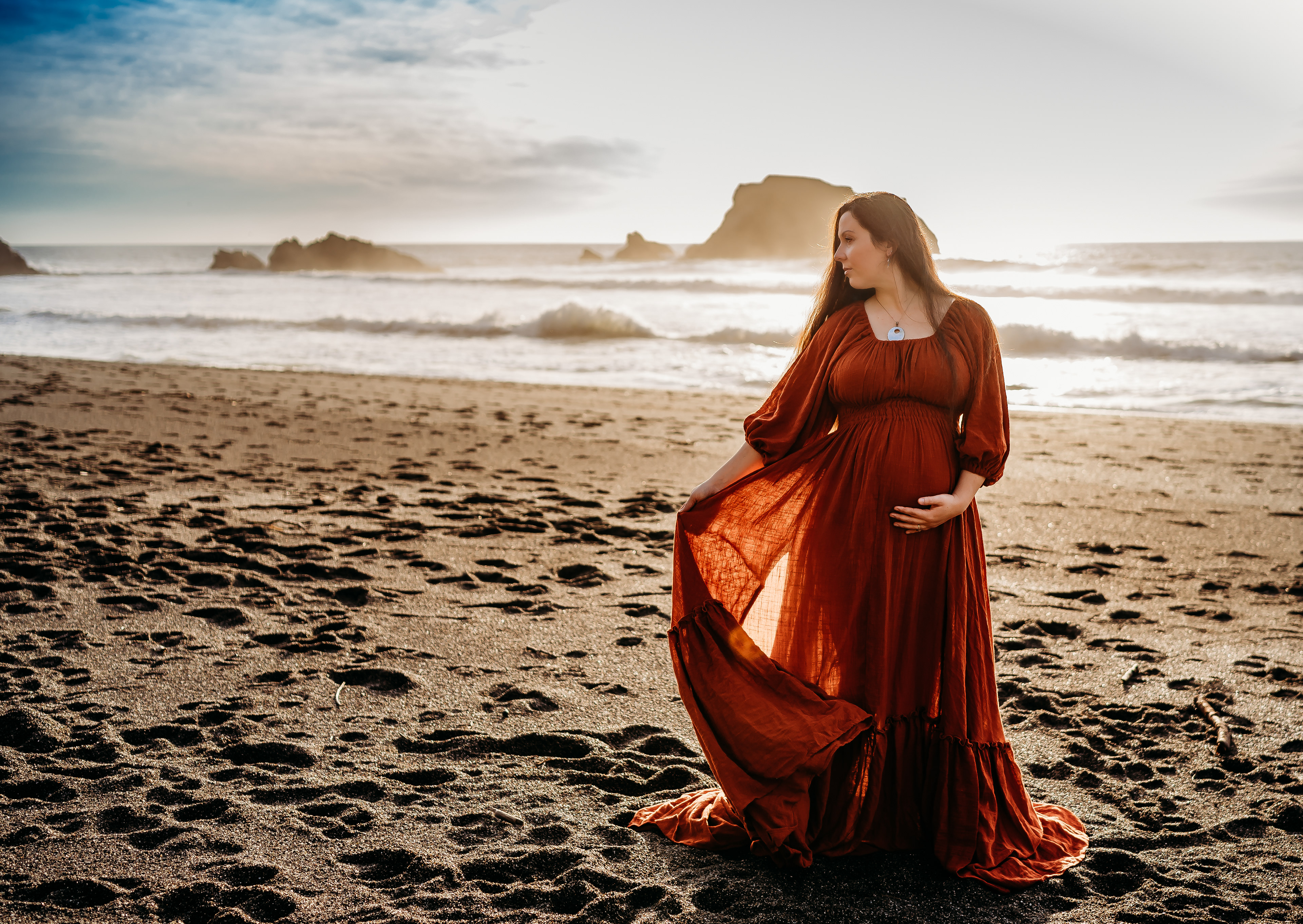 A pregnant brunet women standing on Goat Rock beach in Bodega Bay, California. She is wearing an orange linen dress. She waves the dress in the breeze as waves break on the beach behind her. The picture is taken during a maturnity session with Hearts and Arrow Photography.