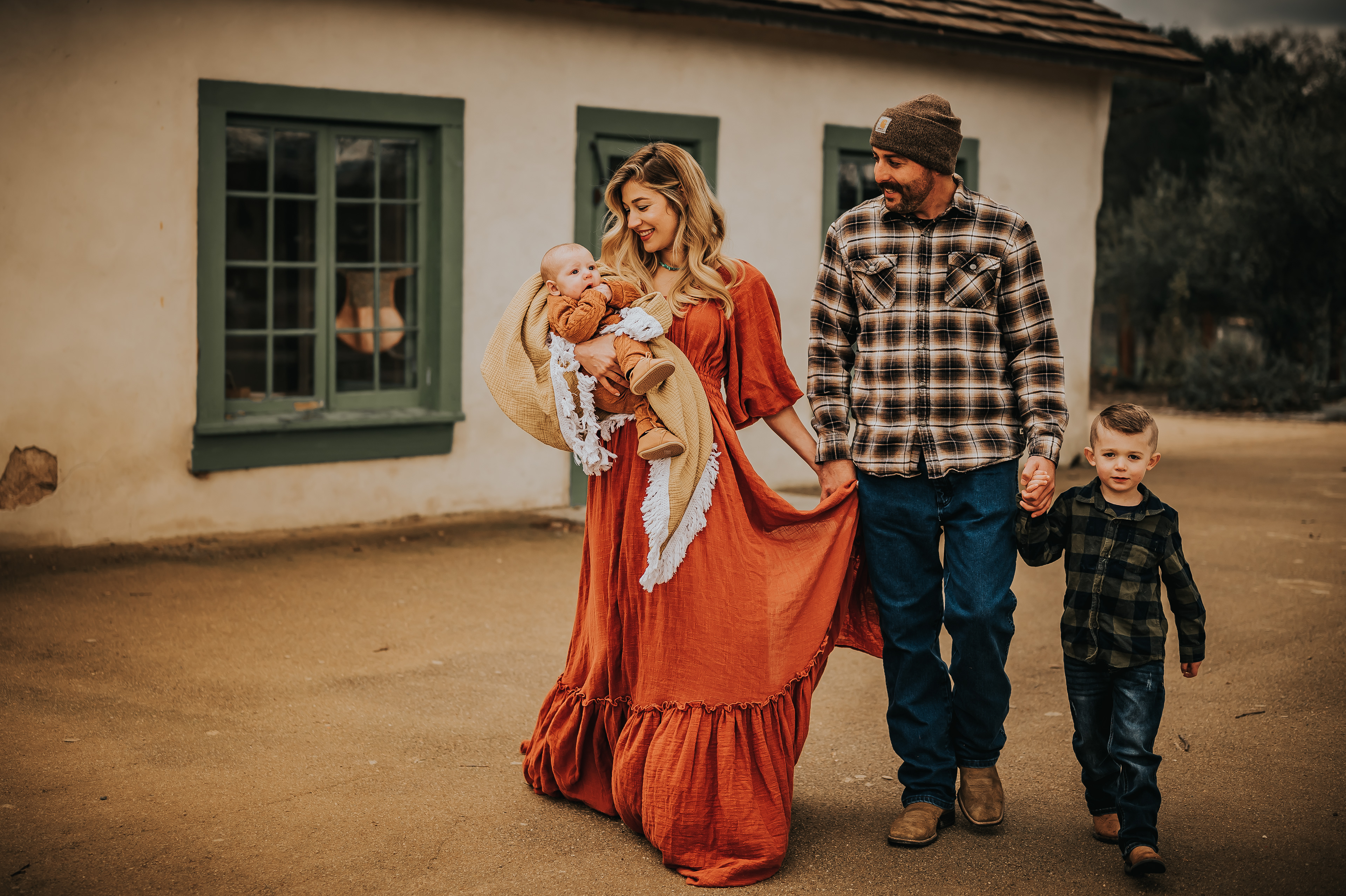 A family of four walking away from an old farm building. Mother is holding a baby in an orange onsie. Father and son hold hands while wearing plaid long sleeve shirts. Photograph taken by Hearts and Arrow Photography in Alviso Adobe Community Park Livermore, California.