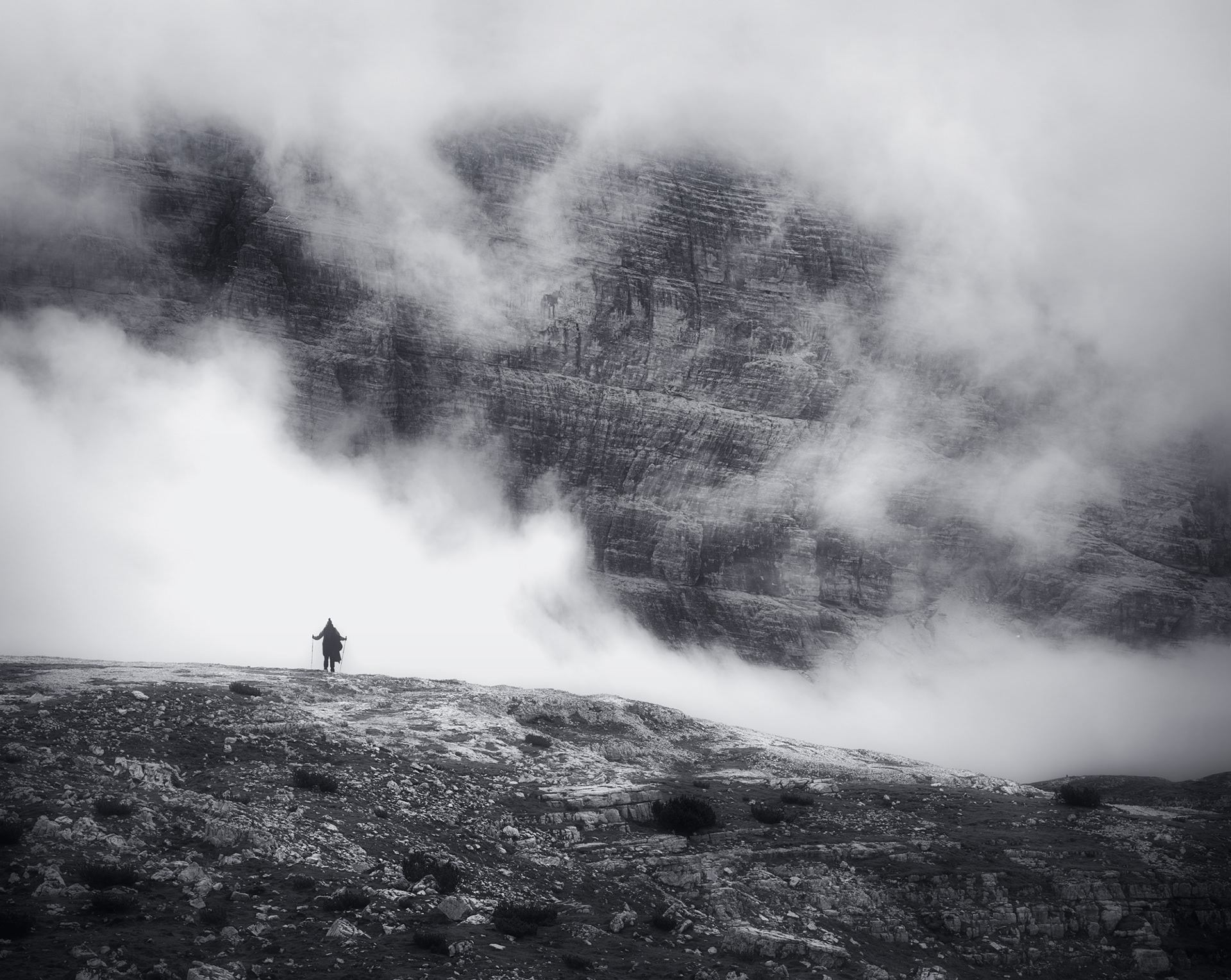 Eine Schwarz-Weiß-Fotografie eines einsamen Wanderers, der auf felsigem Gelände steht, umgeben von dichtem Nebel und Wolken, mit einer dramatischen Bergkulisse im Hintergrund.