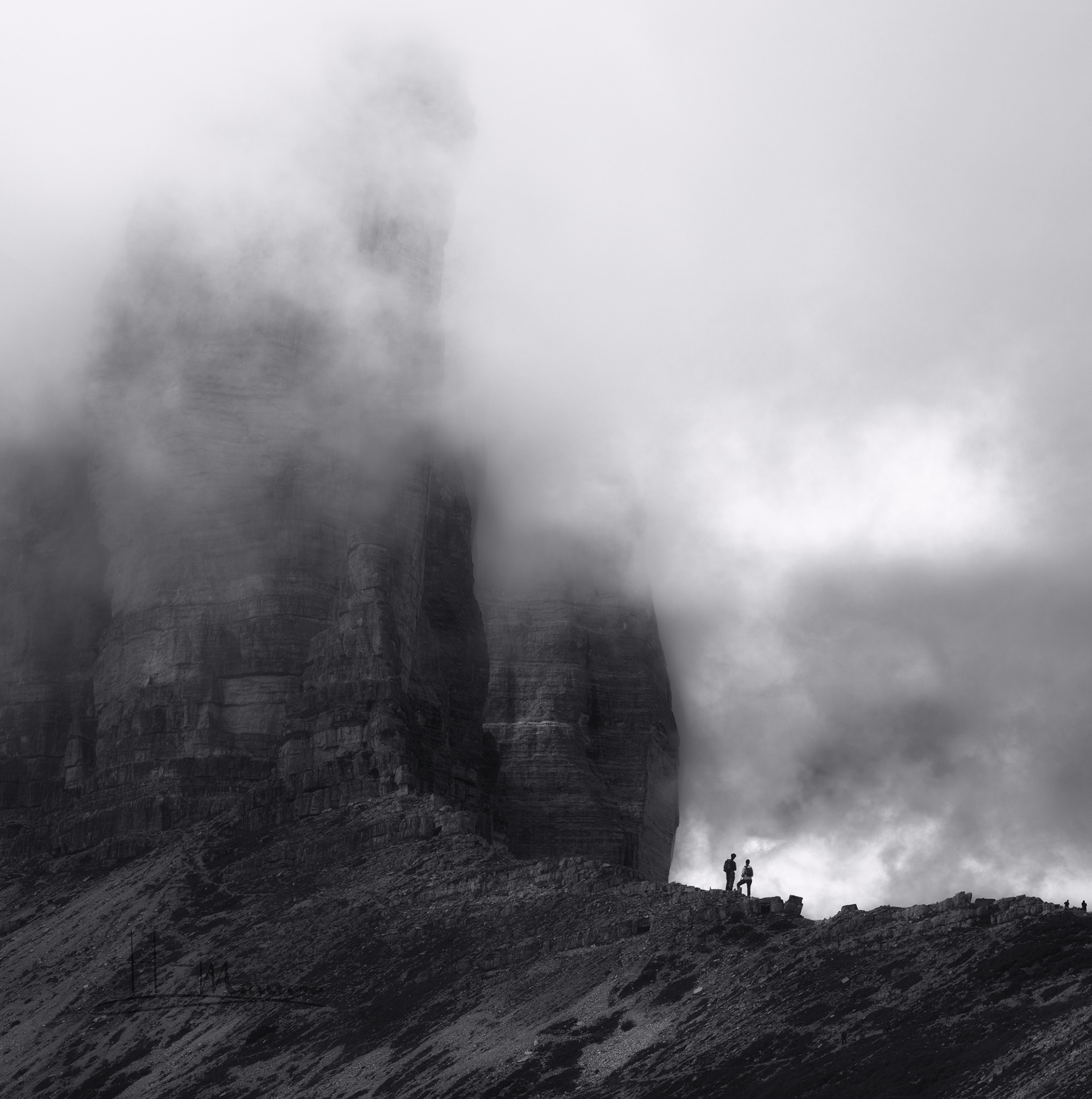 Zwei Wanderer stehen vor einem riesigen, in Nebel gehüllten Berg. Die Landschaft wirkt mächtig und geheimnisvoll, während der Nebel die Klippen teilweise verdeckt. Die Menschen erscheinen winzig im Vergleich zur Natur.