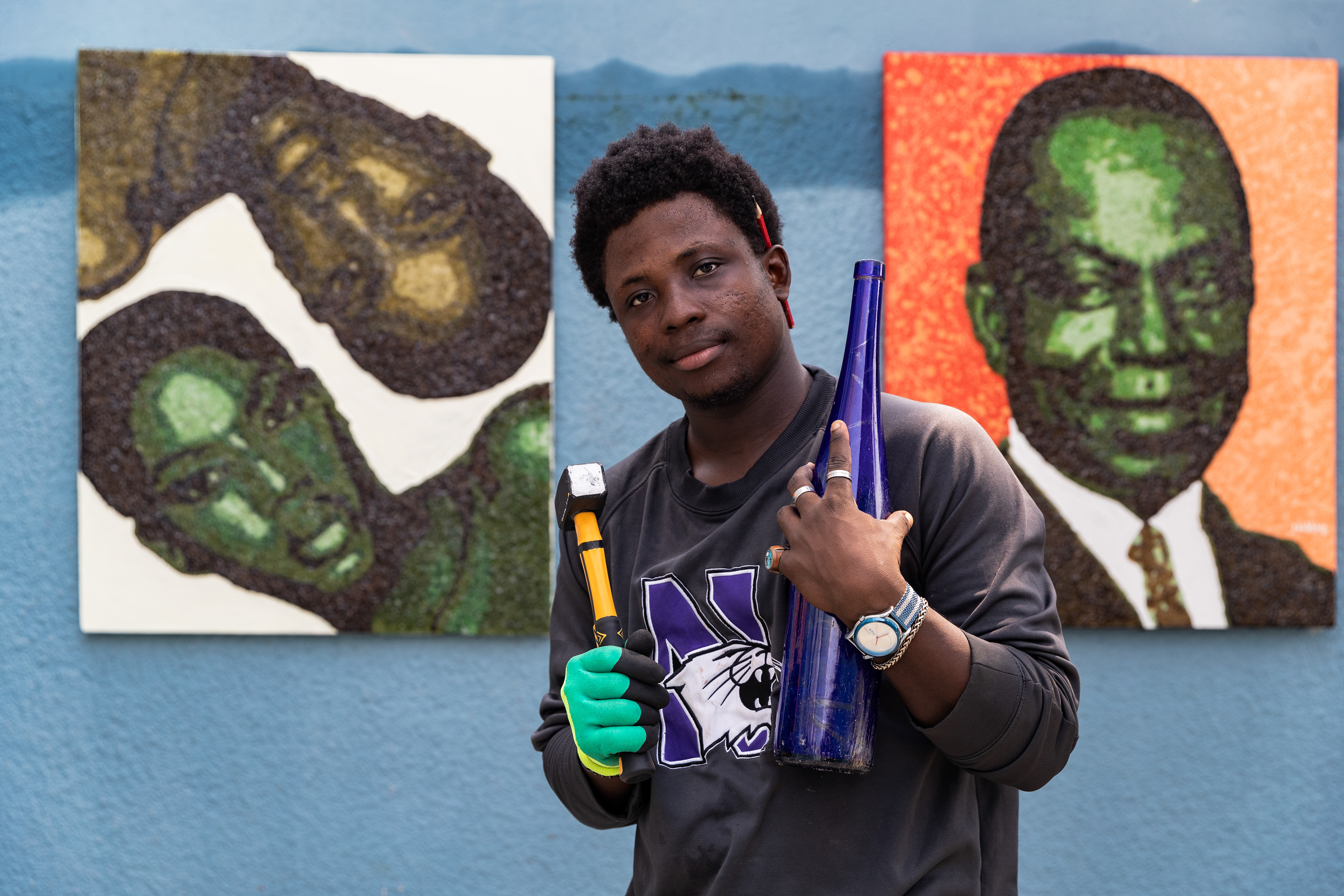 Jonas Doubbin holding a hammer and glass bottle in front of his glass and cigarette collage artworks in his Yopougon atelier, Abidjan, on January 9, 2024, captured by Miléquêm Diarassouba, Photojournalist West Africa.