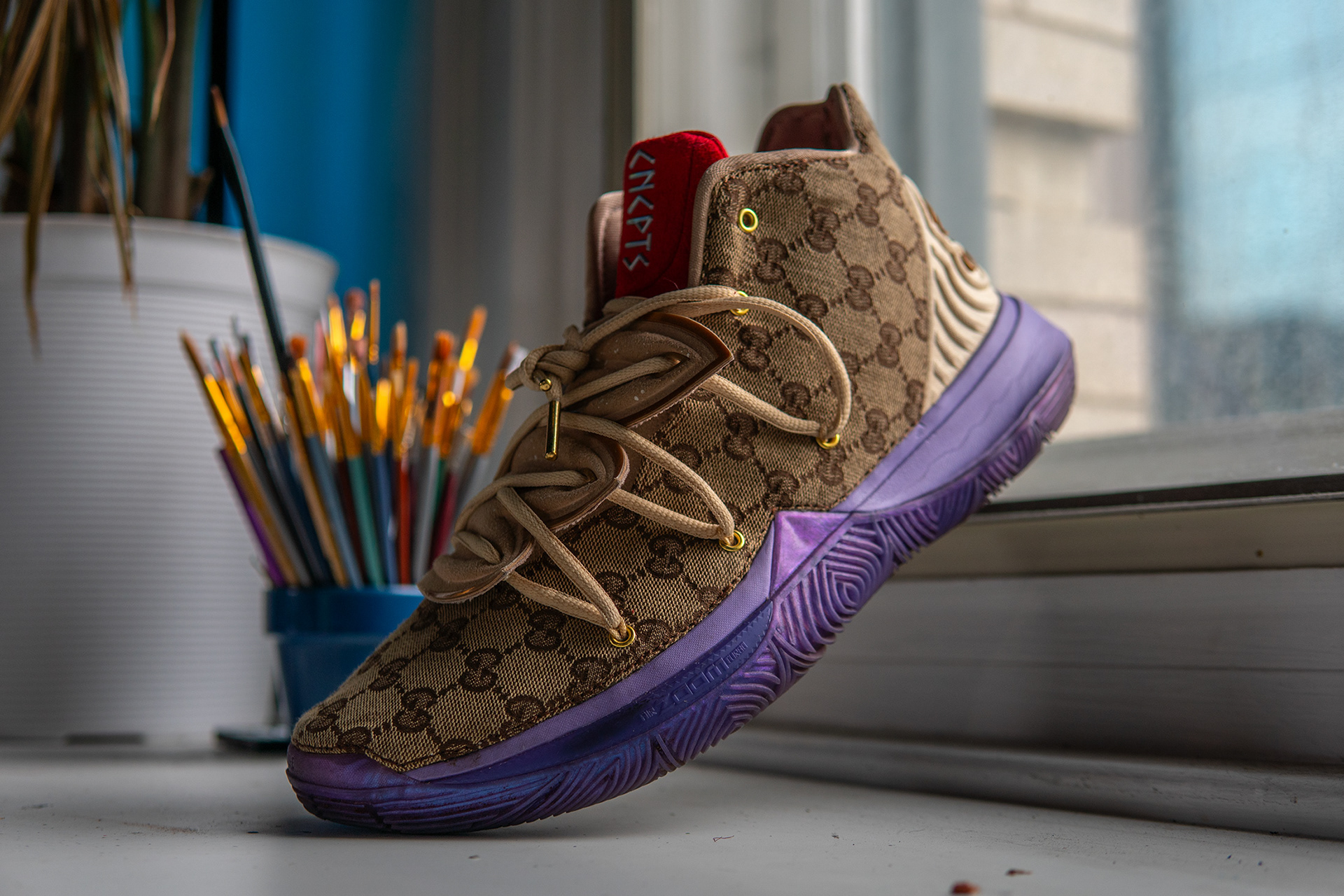 Custom Gucci Nike Kyrie 5 "Ikhet" leaning on a window sill, blurred out paint brushes and planter in the background