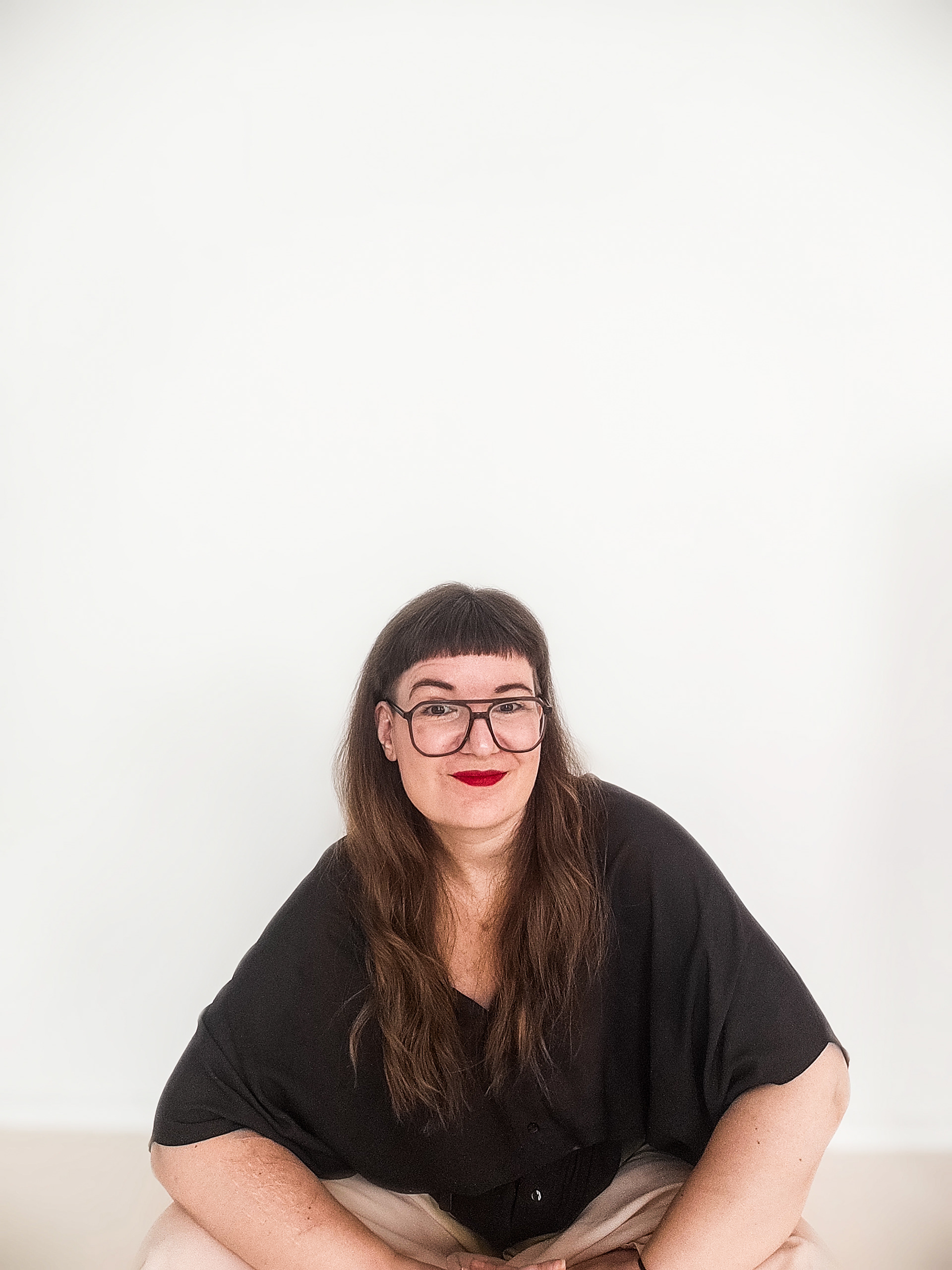 Portraitaufnahme mit weißem Hintergrund sitzend Frau mit schwarzer Brille und schwarzer Bluse und roten Lippenstift.