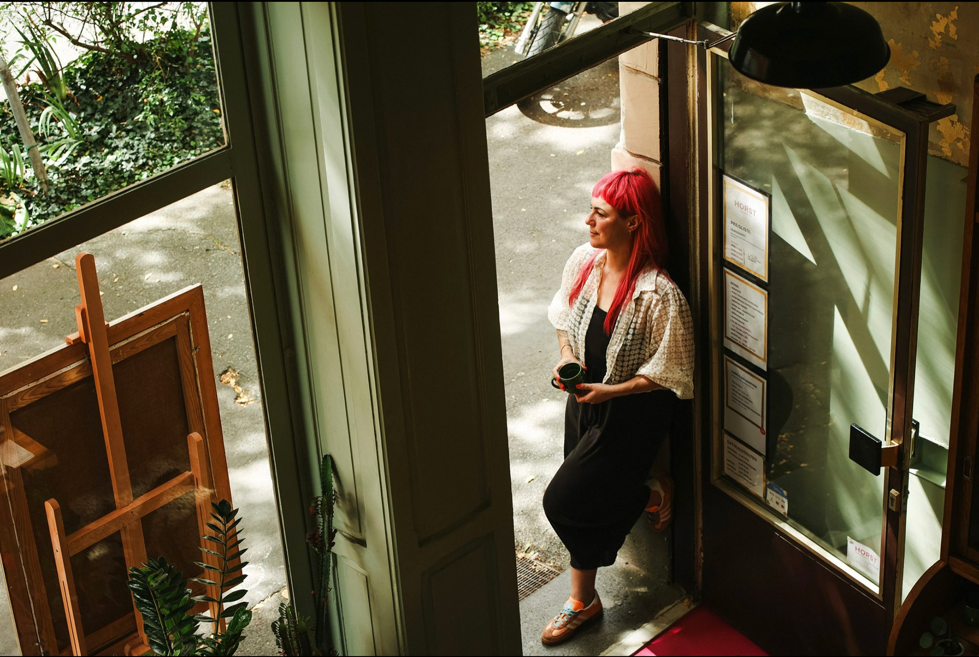 Perspektivische Aufnahme mit Aufsicht im Innenraum auf den Eingangsbereich. Inhaberin mit pinken Haaren und Kaffeetasse im Türrahmen lehnend und auf Straße blickend.