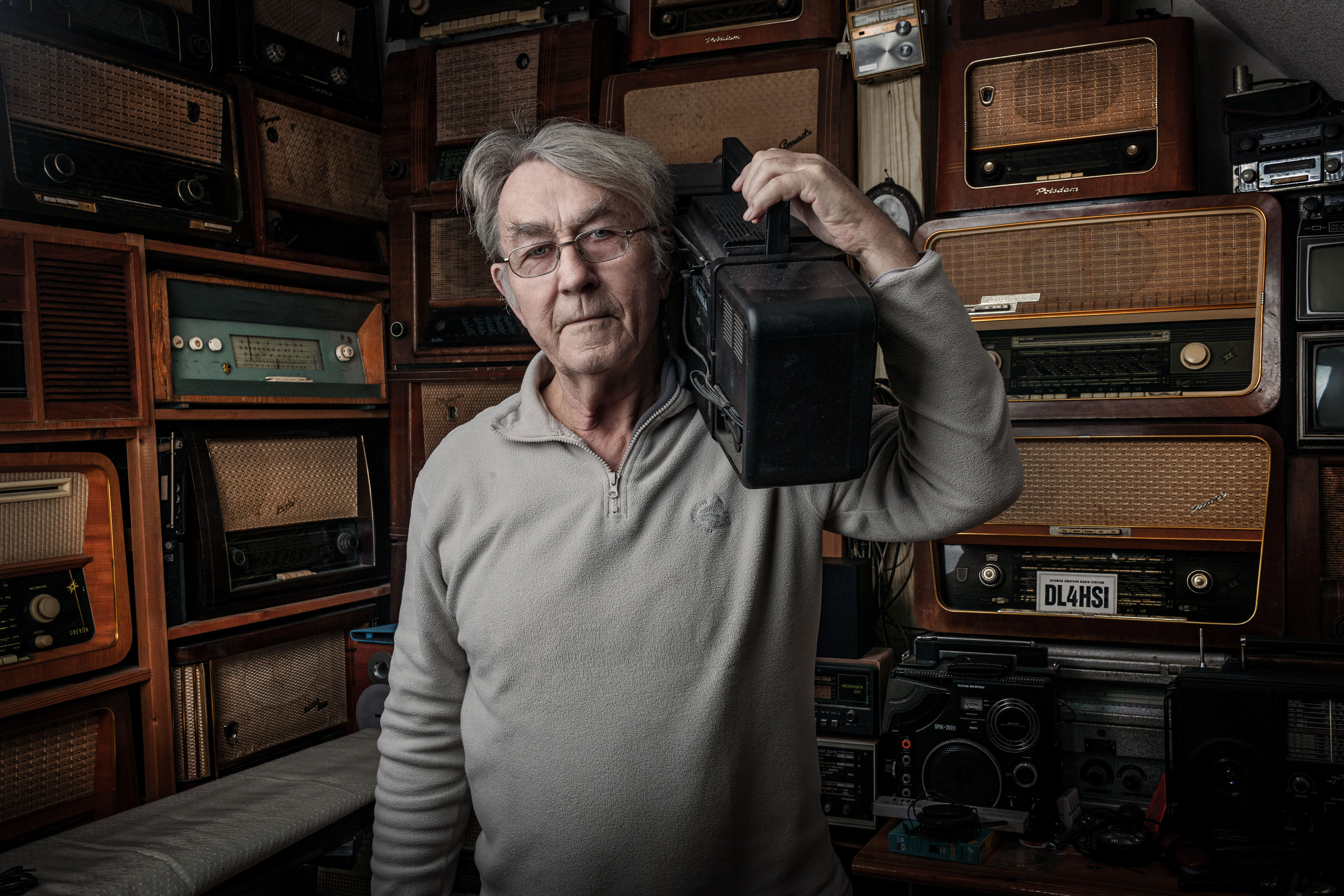 Portrait eines Mannes umgeben von alten Radiogeräten. Er trägt ein Gerät auf der Schulter.