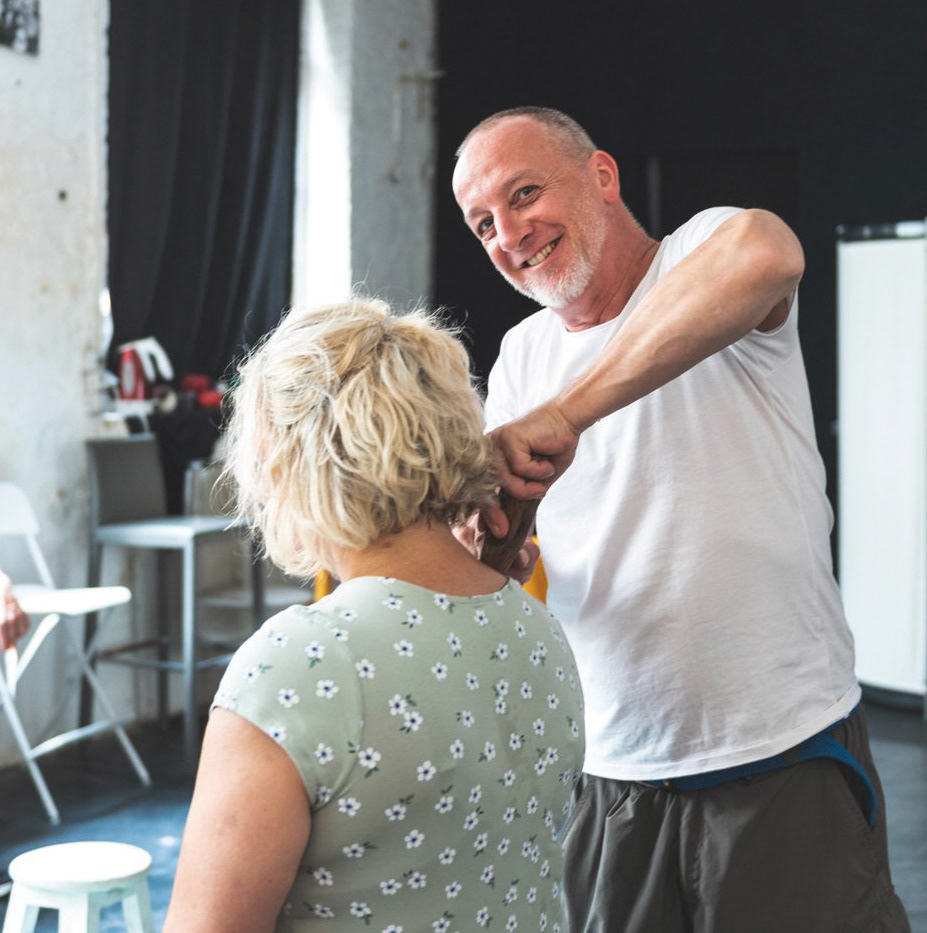 Aufnahme einer Massagesituation mit Faszienholz. Masseur blickt in die Kamera, massierte Frau sitzt mit Rücken zur Kamera.