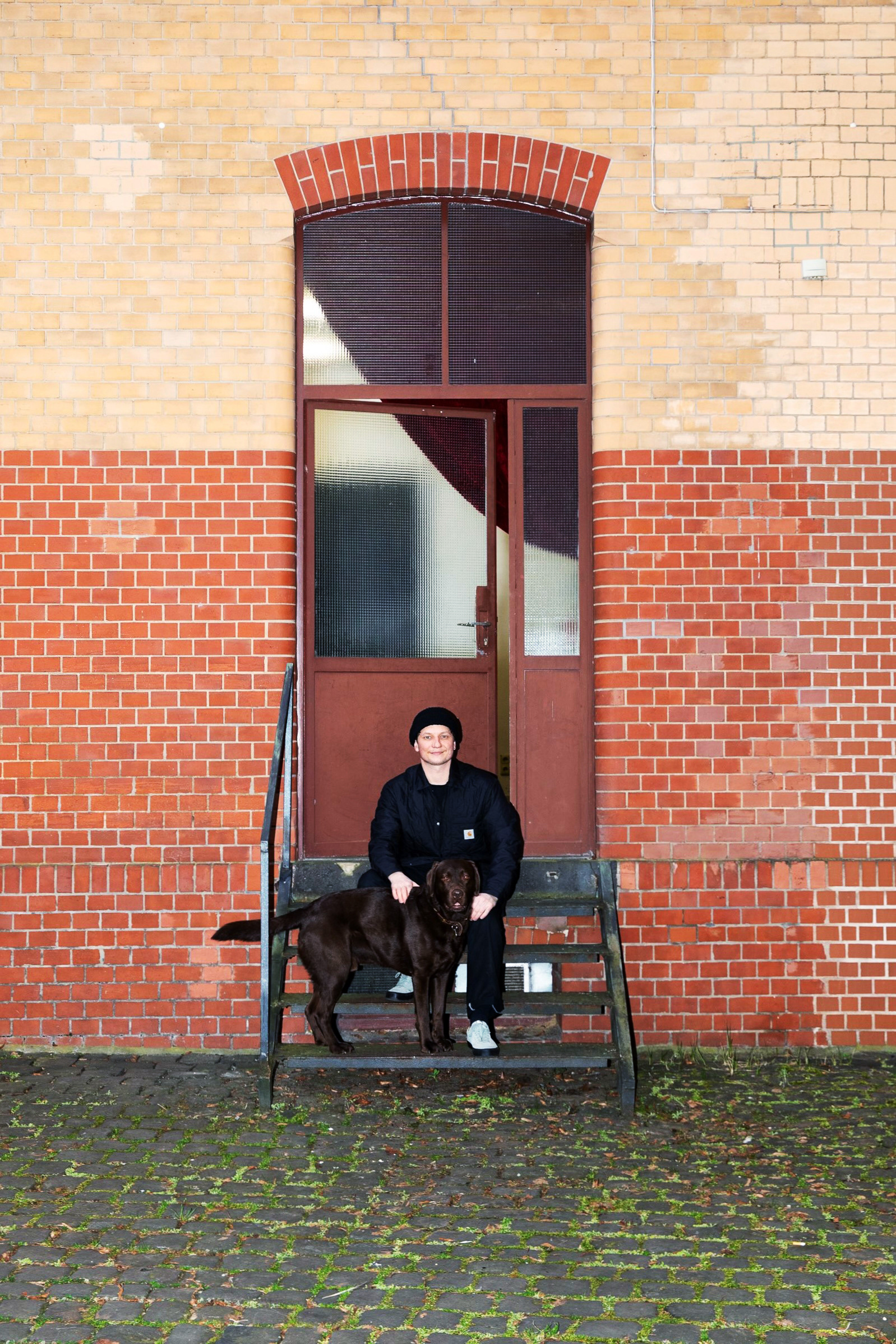 Künstler mit braunen Labrador vorm Eingangsbereich der Werkstatt sitzend auf der Metallzugangstreppe. Backsteinfassade.