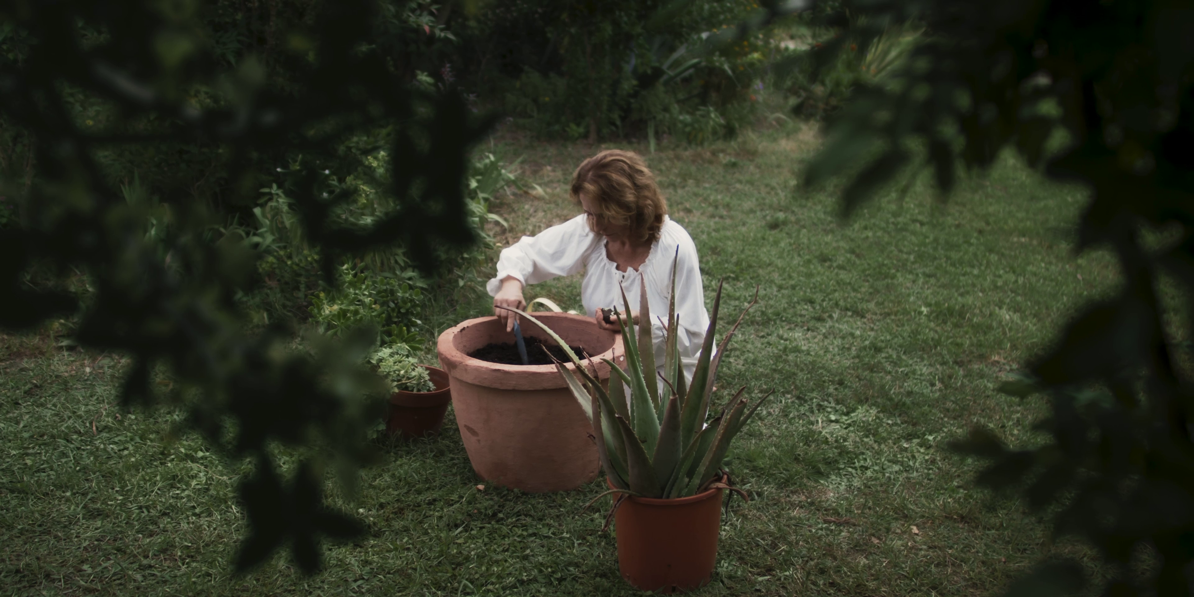 Ein Filmstill aus dem Film "EDEN". Die Protagonistin Sitzt Neben einem Großen Topf in auf einer Wiese und Pflanz etwas in ihm ein.