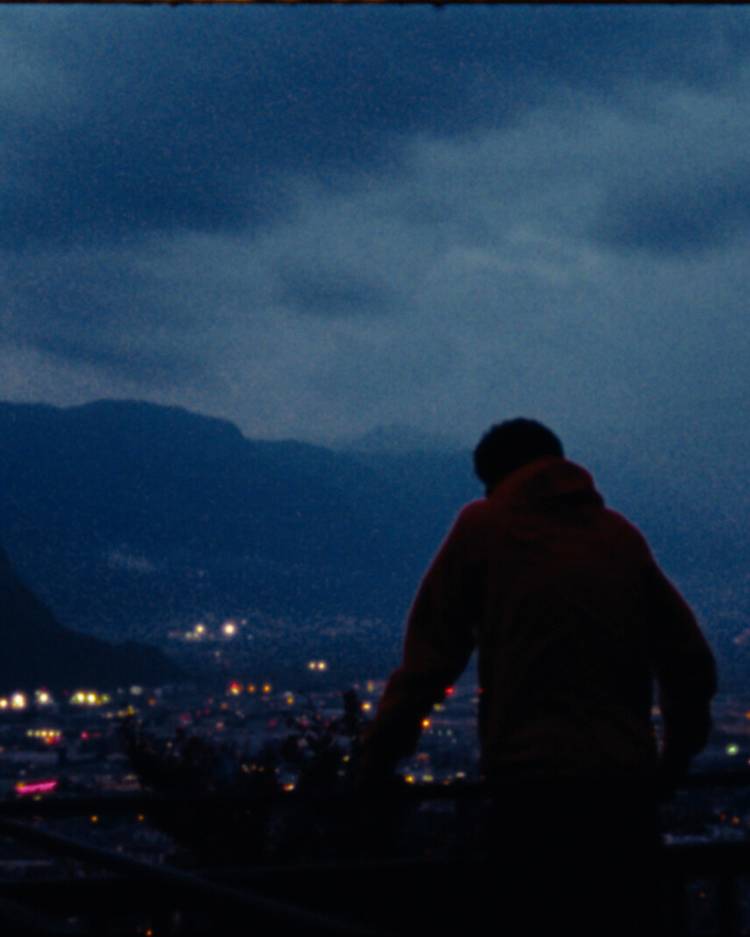 Ein Filmstill aus dem Film "TSHEGL". Ein Junger Mann schaut im Abendlicht, von Weiter oben, auf eine Mit Bergen umringte Stadt, in der Lichter leuchten.