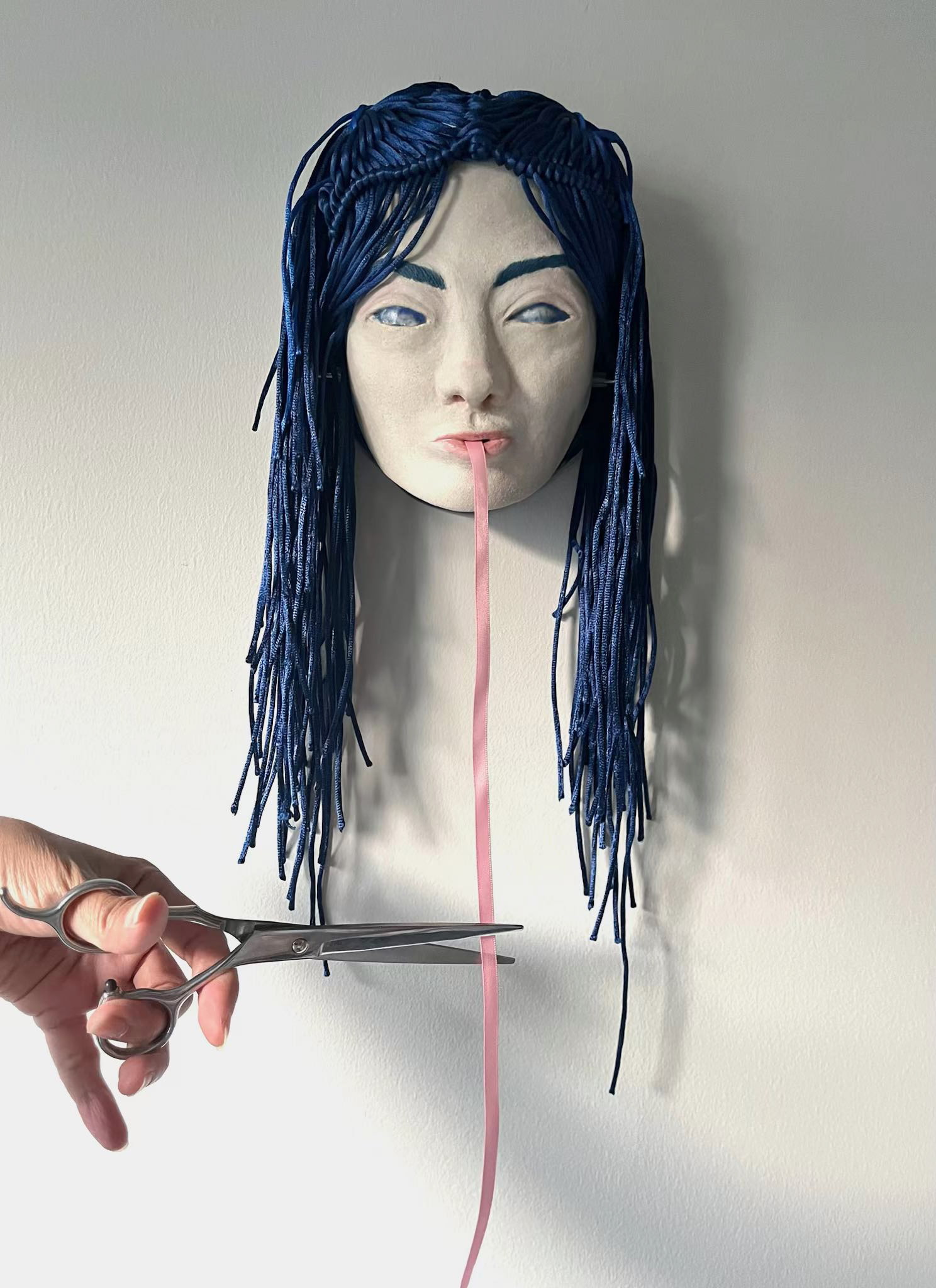 The Tiktik is a shape-shifting monster from Philippine folklore. It sneaks onto rooftops and lowers a thin long tongue to consume its sleeping victims. It can be repelled by garlic or a pair of open scissors.  My piece invites the viewer to cut off a piece of the tongue, a long continuous ribbon protruding from its mouth.  Ceramic sculpture, knotted cord, and ribbon. A spool mechanism hidden within the face.