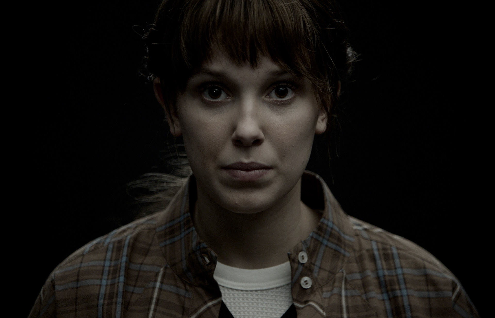 Close-up portrait of a Eleven played by Millie Bobbie Brown with bangs in a plaid shirt against a dark background, from the Stranger Things Immersive Experience film