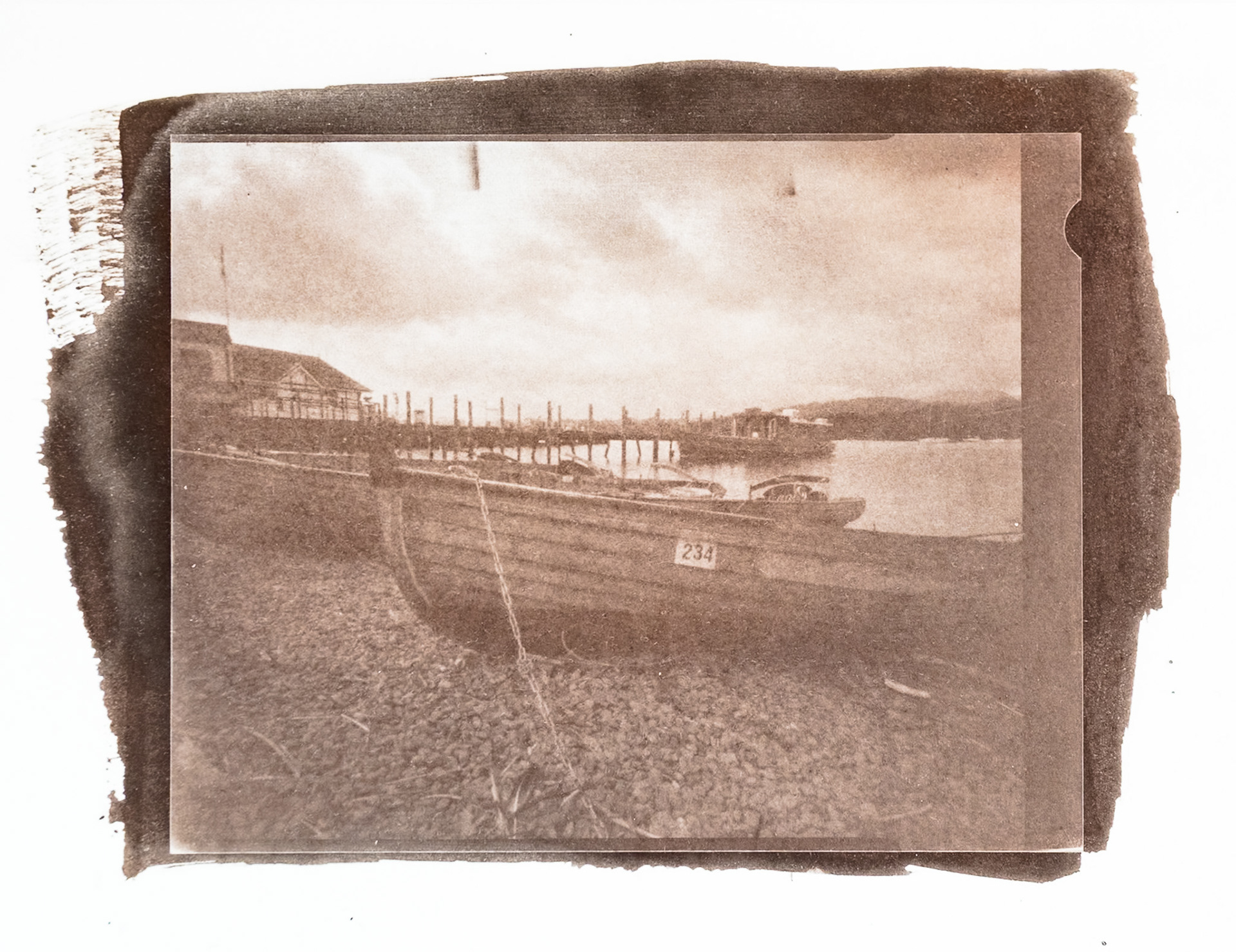 Fine art hand printed argyrotype print of  a row of beached rowing boats on the shore of Lake Windermere. A Jetty and gently rolling hills form the background.