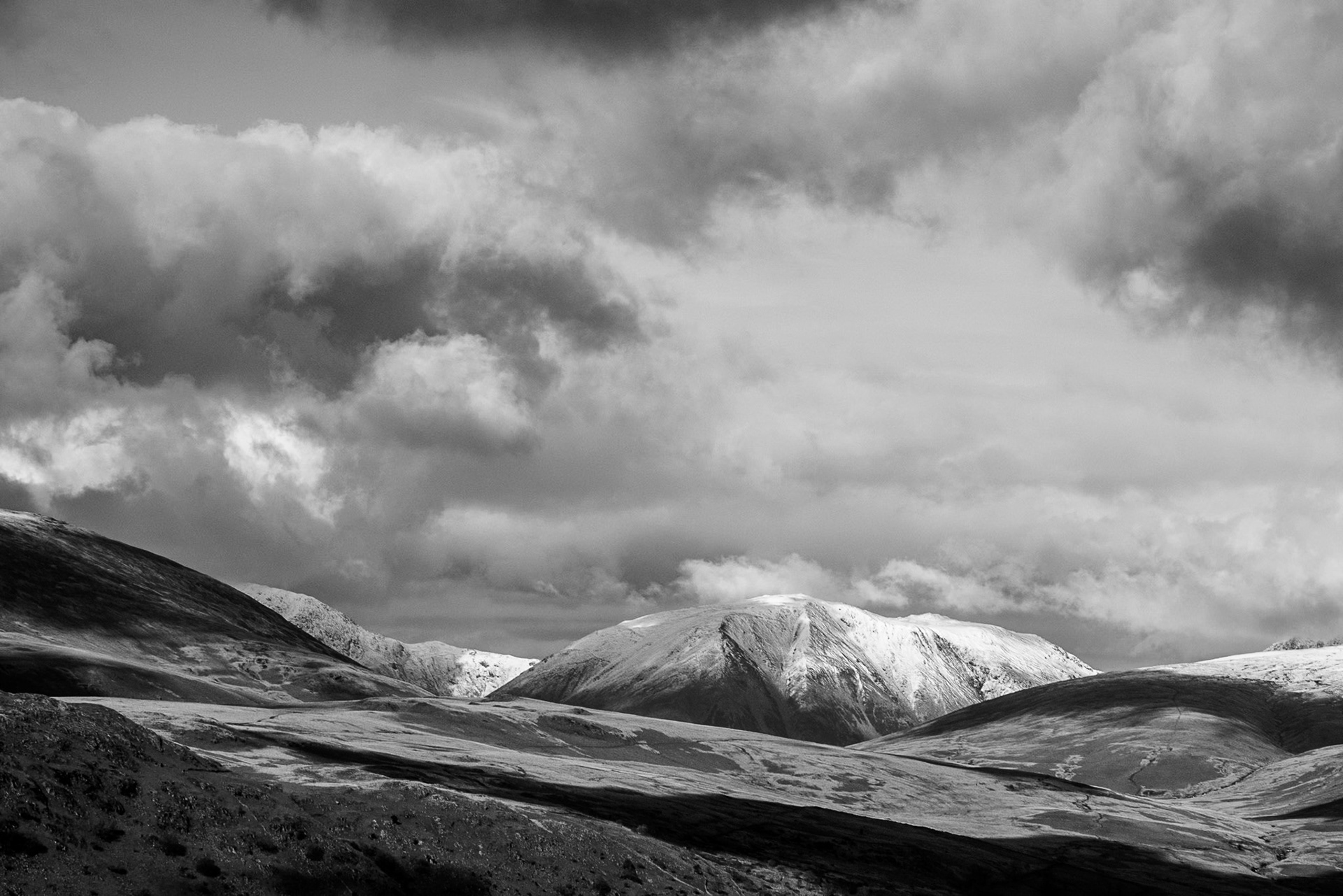 Black-and-white mountain landscape with snow-covered peaks, barren rolling hills, and dramatic storm clouds creating a stark, moody scene.