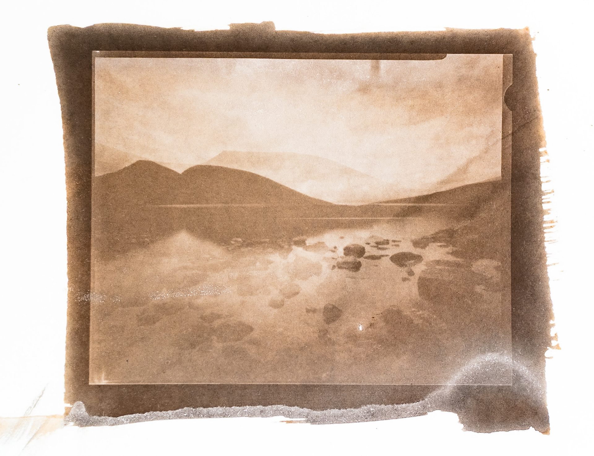 Fine art hand printed argyrotype print of a double exposed negative.  The image shows a rocky lake and rolling hills.