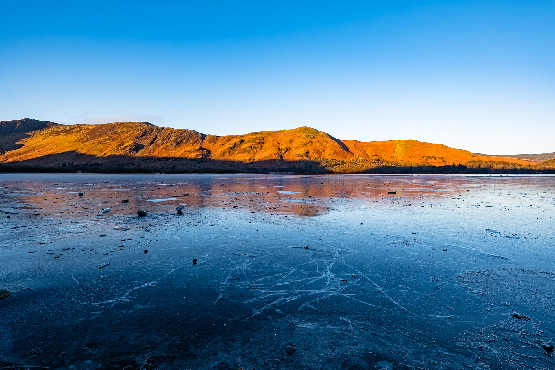 Early morning golden light floods across the fells behind  the frozen shore of Derwent water in Cumbria.