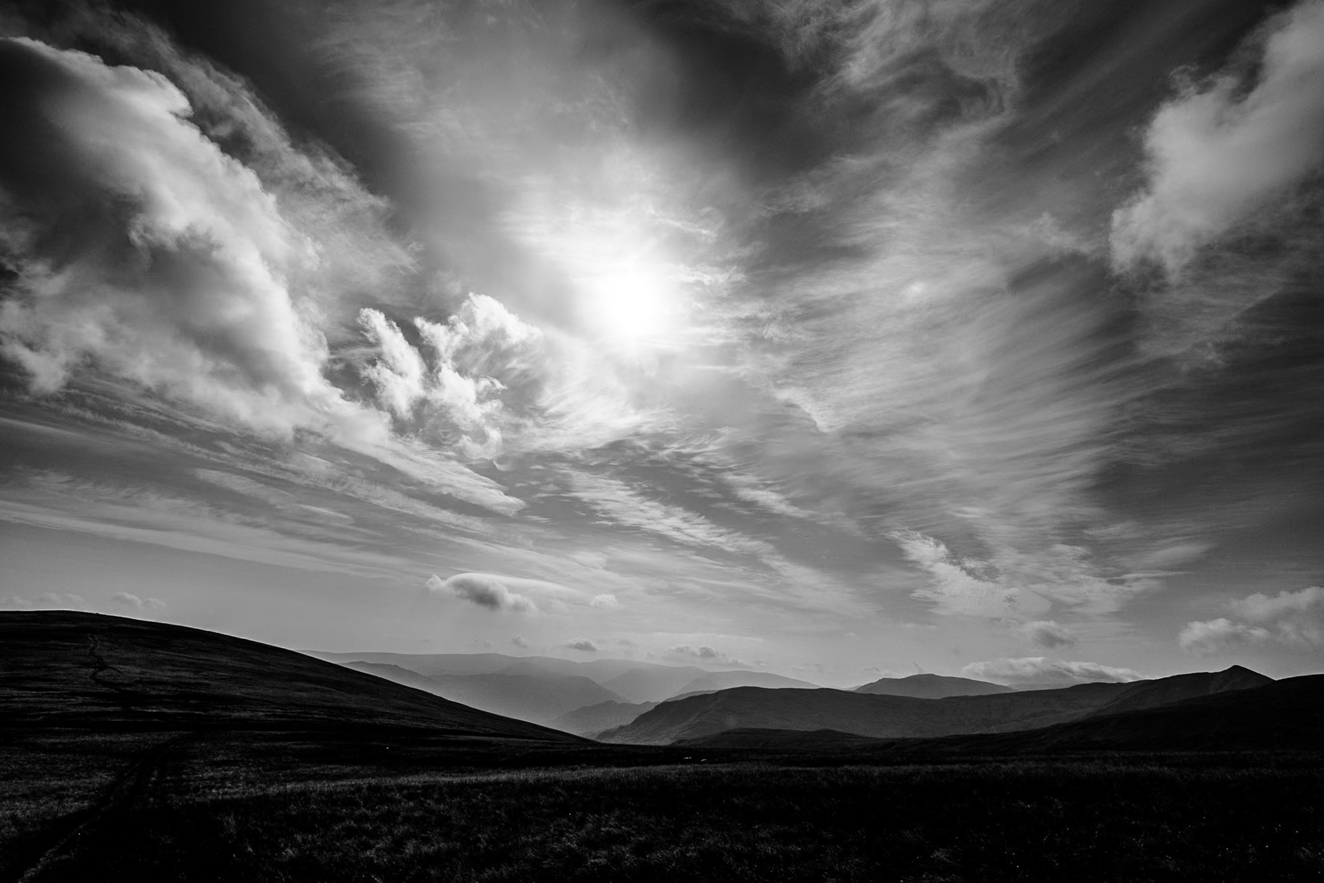 Black-and-white mountain landscape beneath dramatic swirling clouds and bright sunlight, creating a vast, serene yet powerful atmosphere.