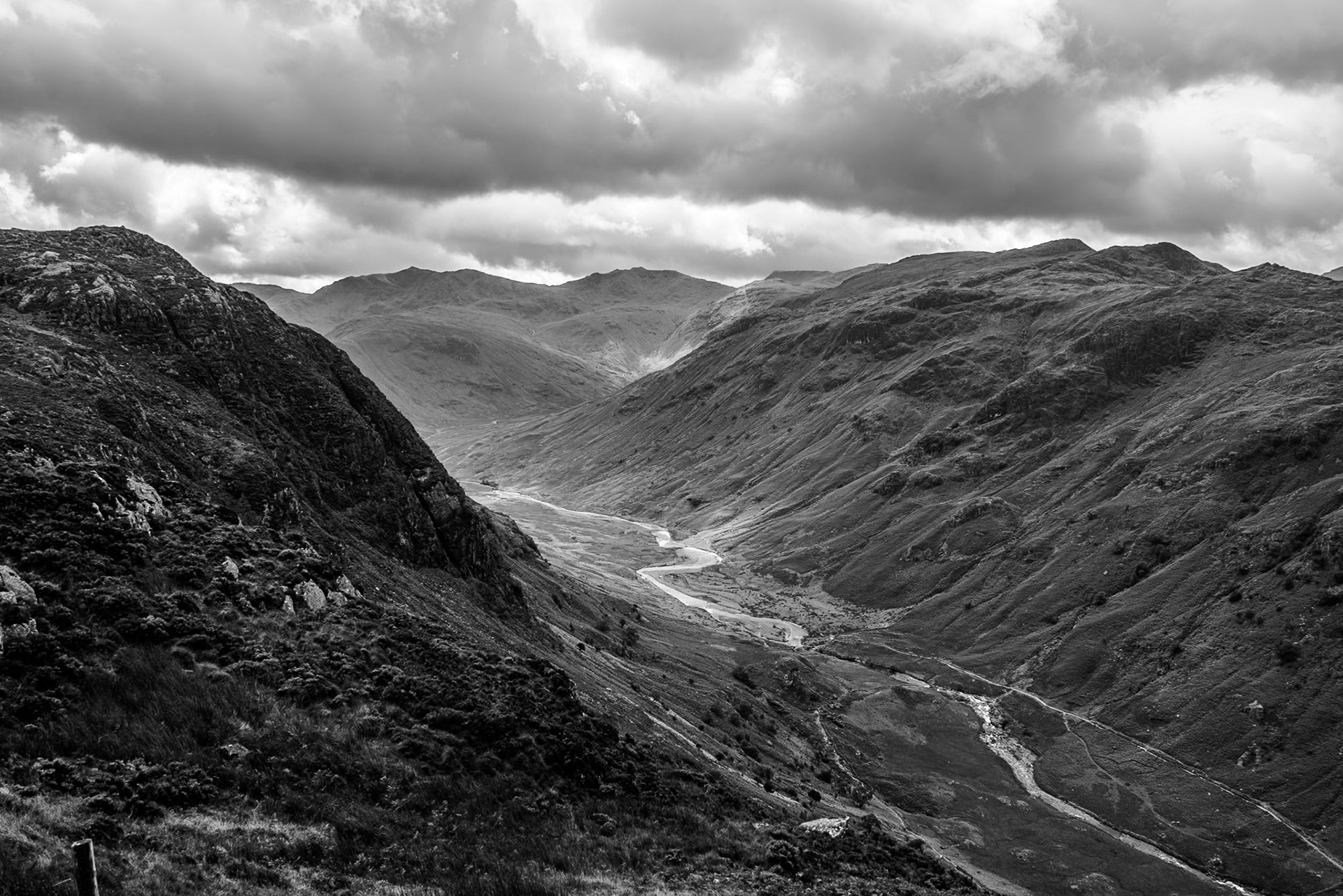 A black and white image of a steep sided and rugged mountain valley with patches of sunlight illuminating a mountain beck. 