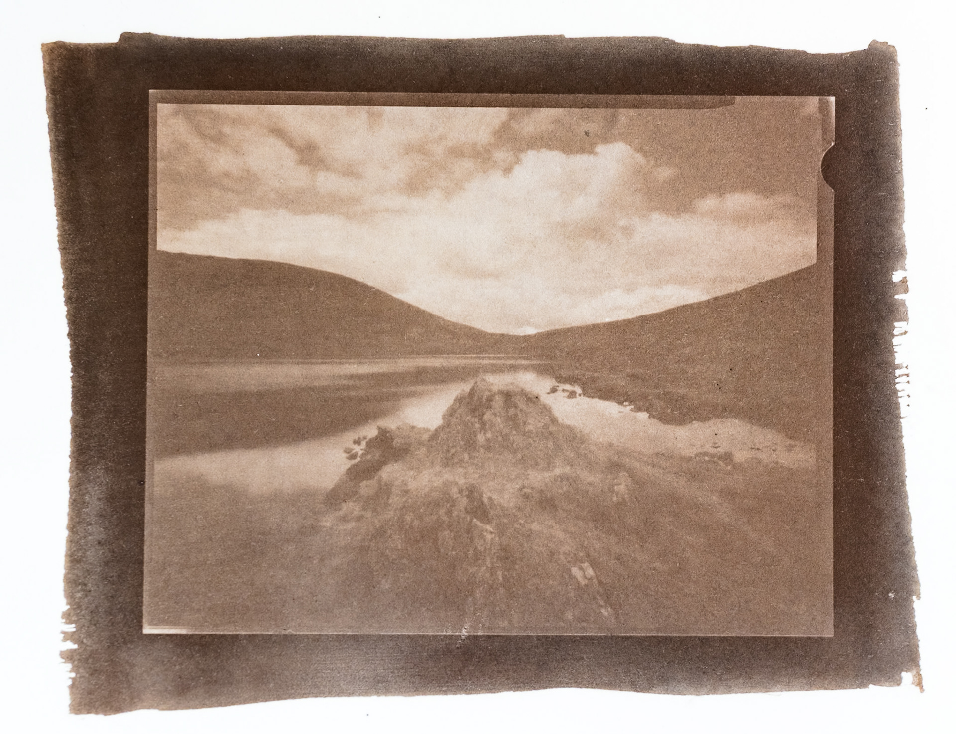 Fine art hand printed argyrotype print of rocks jutting out into a still lakeland tarn.  Rolling hills and billowing clouds can be seen in the background.