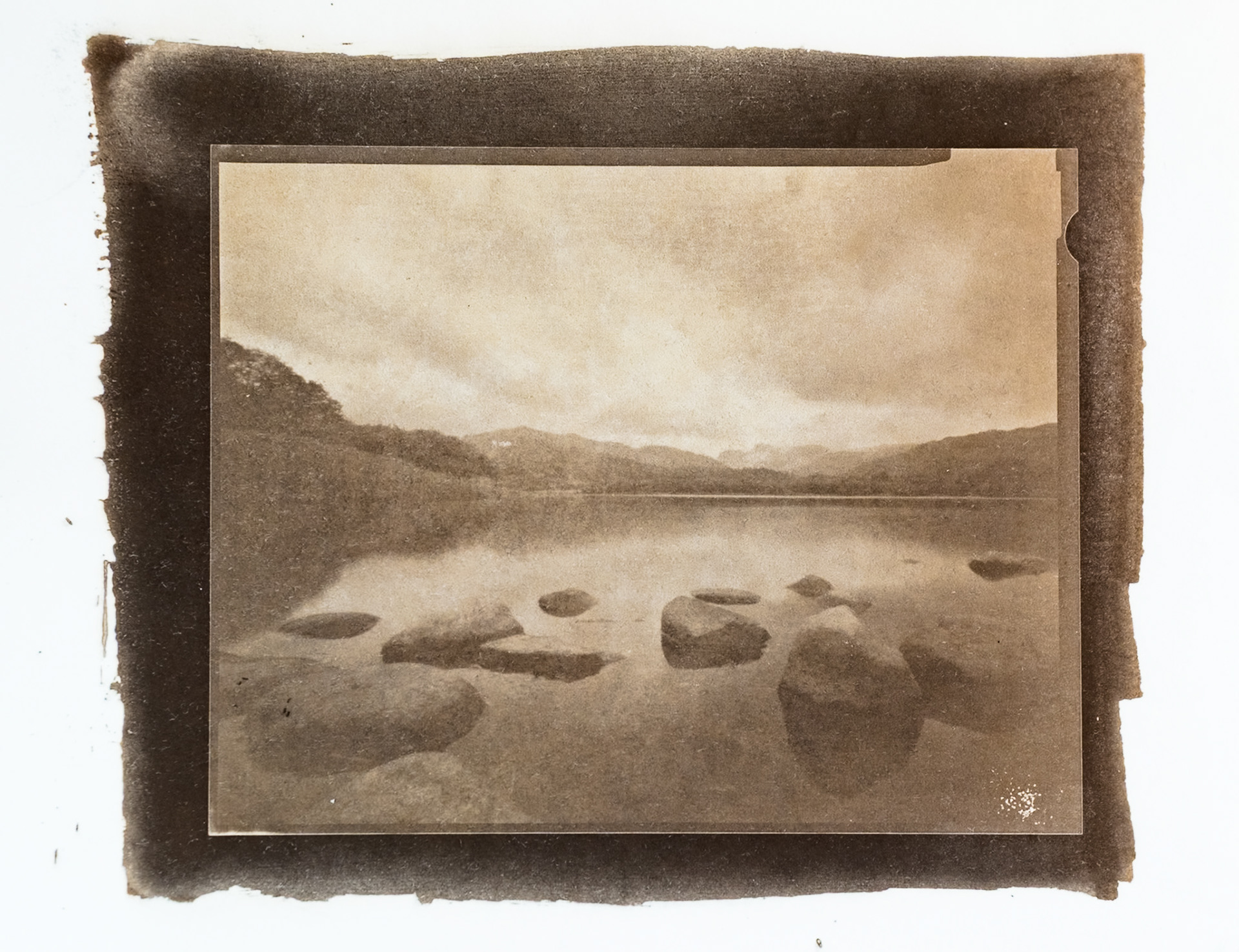 Fine art hand printed argyrotype print of a calm Elterwater Lake. Partially submerged rocks can be seen in the foreground and the Langdale Pikes can be seen in the distance.