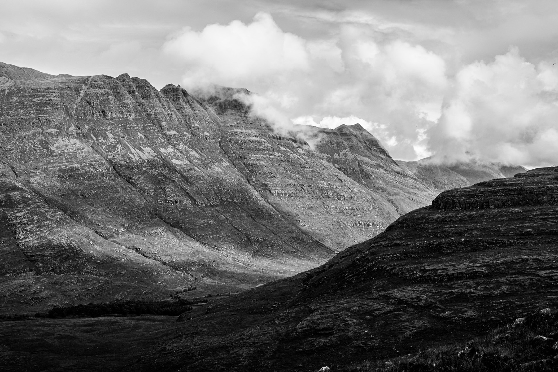 Black-and-white mountain landscape with rugged ridges, a deep valley, and billowing clouds overhead, creating a dramatic, tranquil mood.