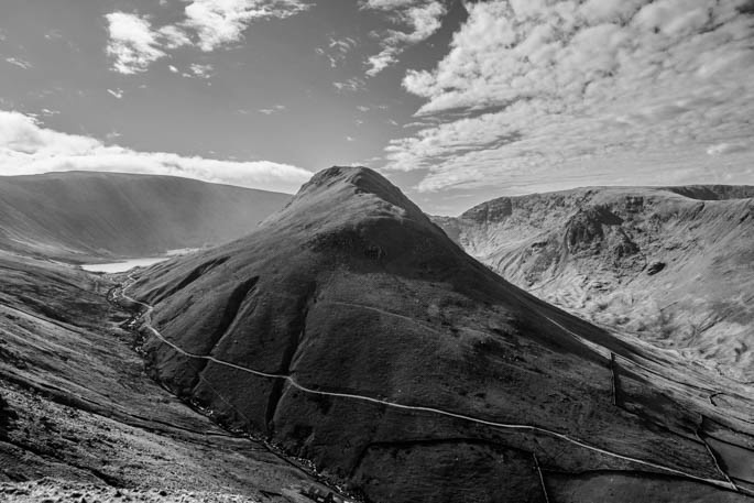 A black and white image of Gray Crag in shadow.  The mountain has a thin track along its base and Haweswater reservoir can be seen in the distance.