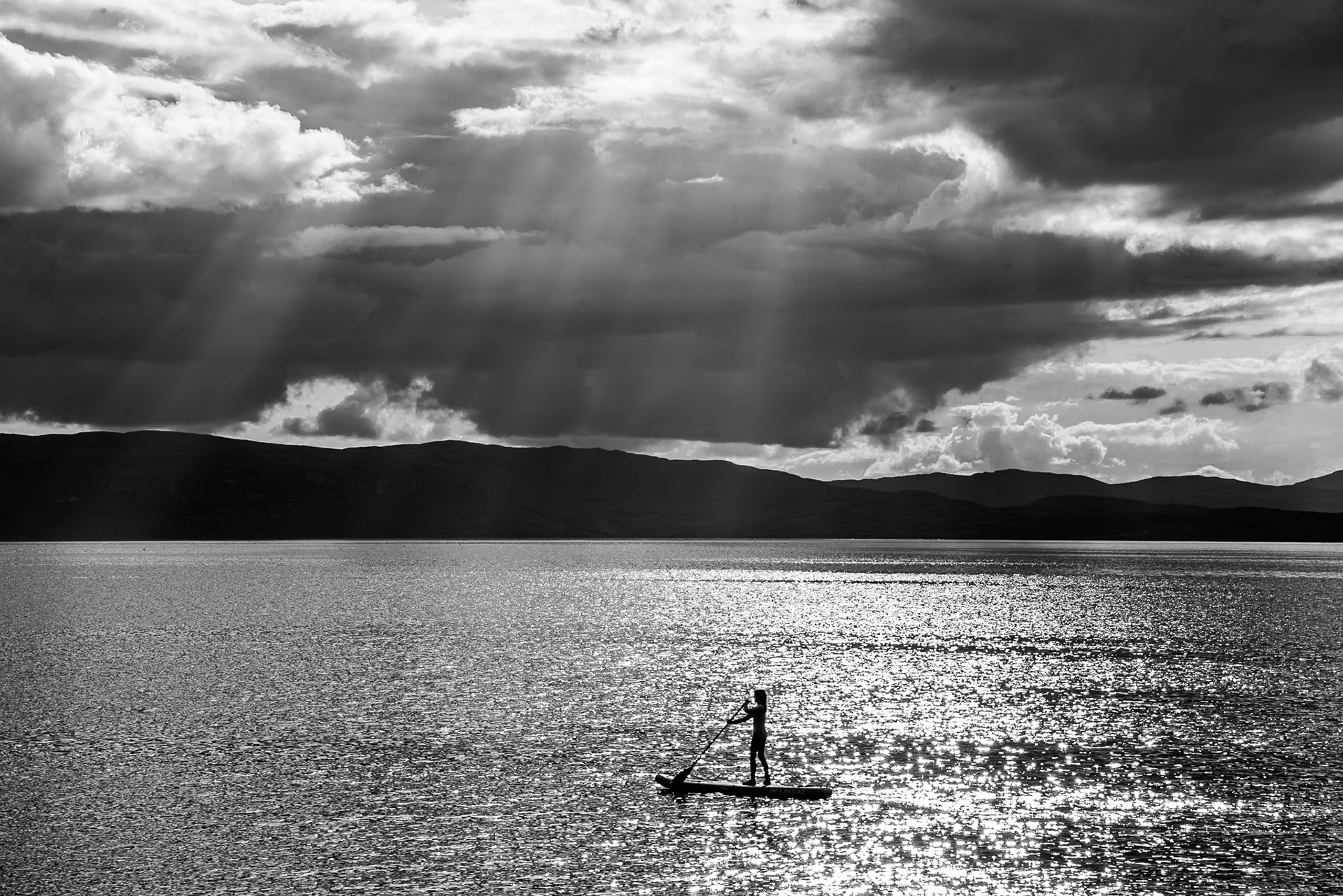 A lone paddleboarder crosses a sunlit Applecross Bay beneath dramatic, dark storm clouds, with a silhouetted mountain ridge stretching across the horizon.