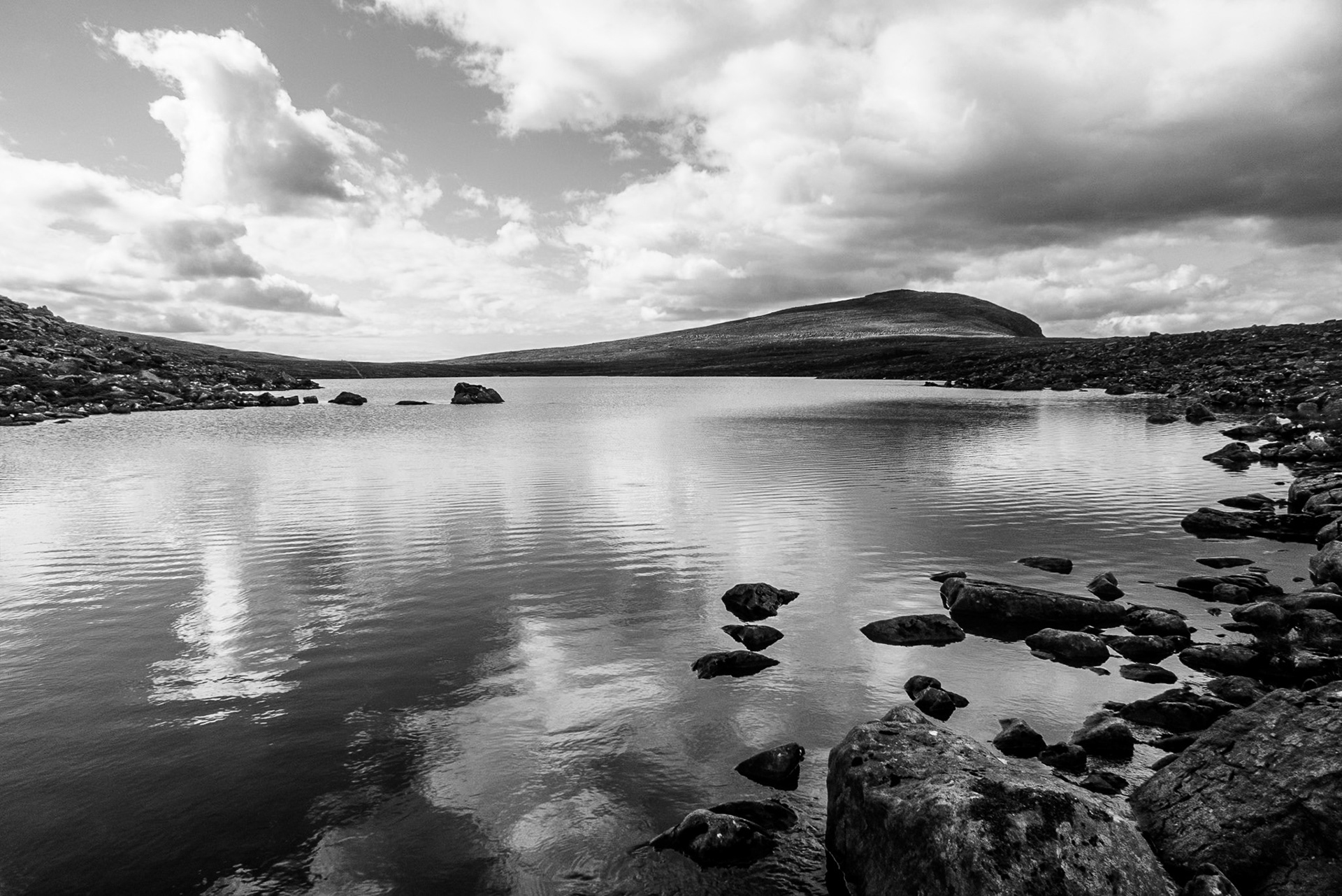 Black-and-white mountain loch high in the Torrid Mountains in North West Scotland with calm reflective water, rocky shoreline and dramatic clouds.
