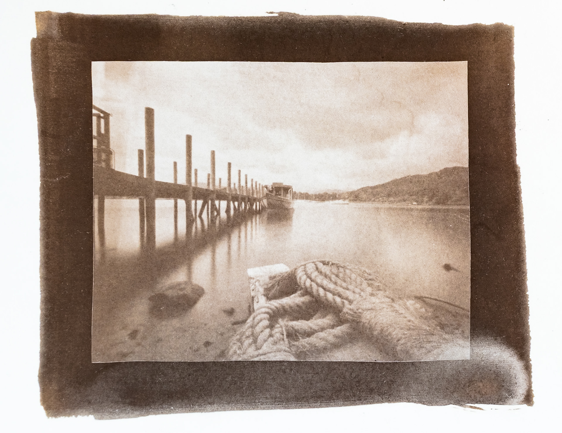 Fine art hand printed argyrotype print of  a jetty stretching out into Lake Windermere.  A Boat is tied up at the jetty and a coiled rope can be seen in the foreground.