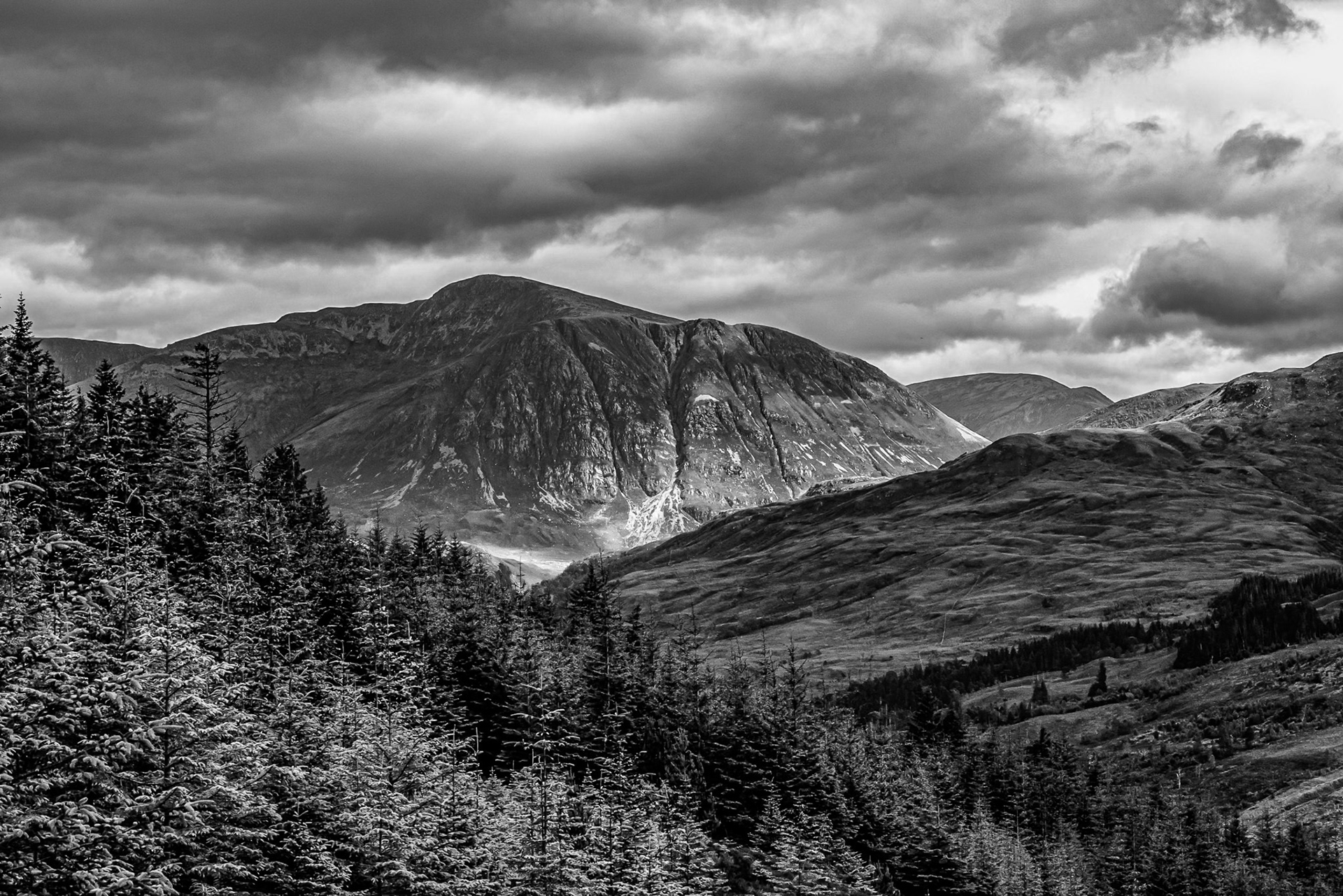 Black-and-white mountain landscape with rugged peaks, dark evergreen forest, rolling slopes, and dramatic storm clouds.