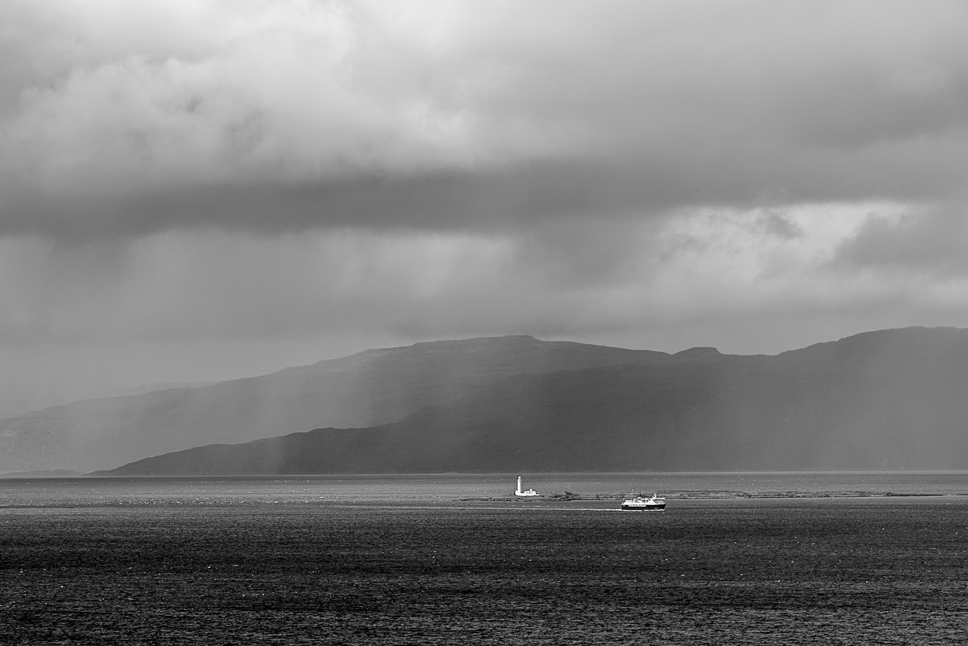 The Oban Ferry passes the Eilean  Musdile lighthouse, with rain swept mountains and a cloudy sky creating a dramatic, atmospheric scene.
