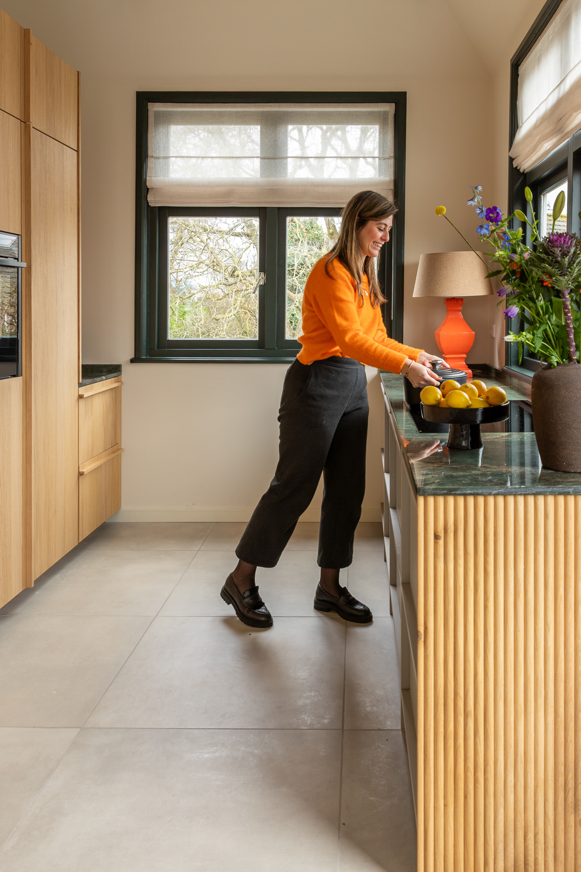 interieur houten keuken met groen werkblad, grijze tegel vloer en groene kozijnen, met vrouw in oranje trui