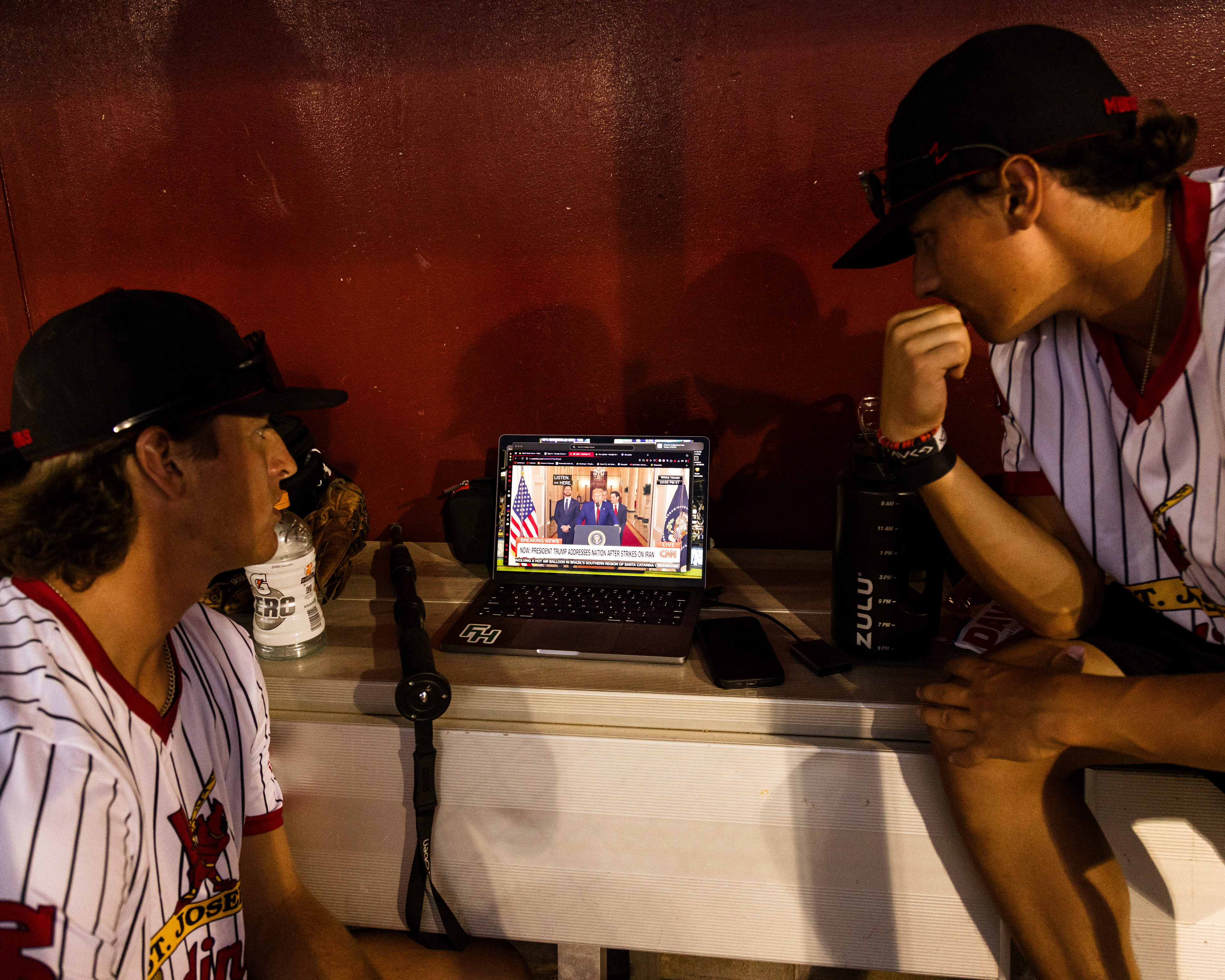From left, Aron Harrington and Sam Pitts watch Donald Trump announce the bombing of Iran on a laptop in the dugout 