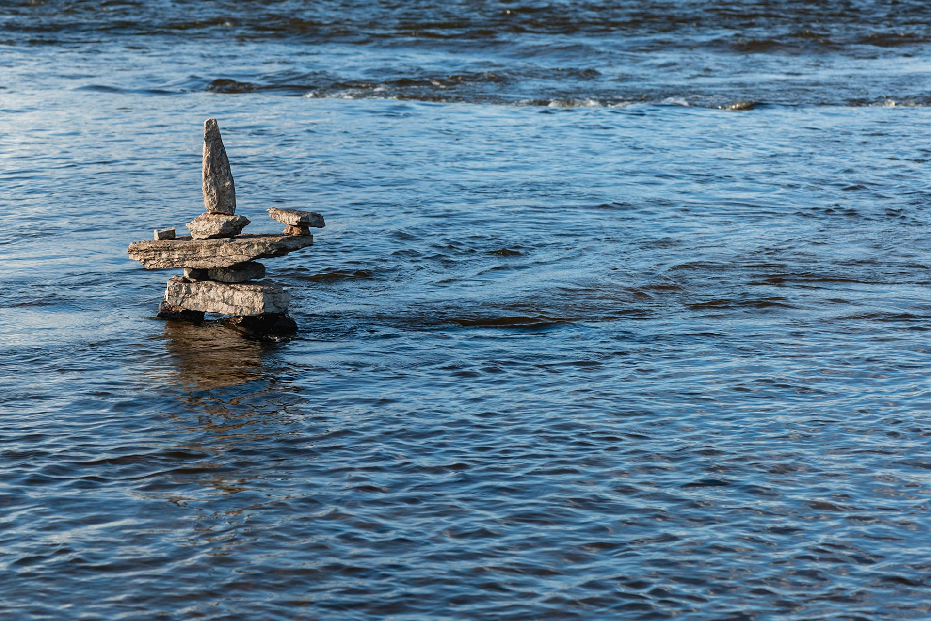 An inukshuk sculpture in the river. Turbulent water at the top of the frame, along with the wake of the sculpture, give a sense of the speed of the water.