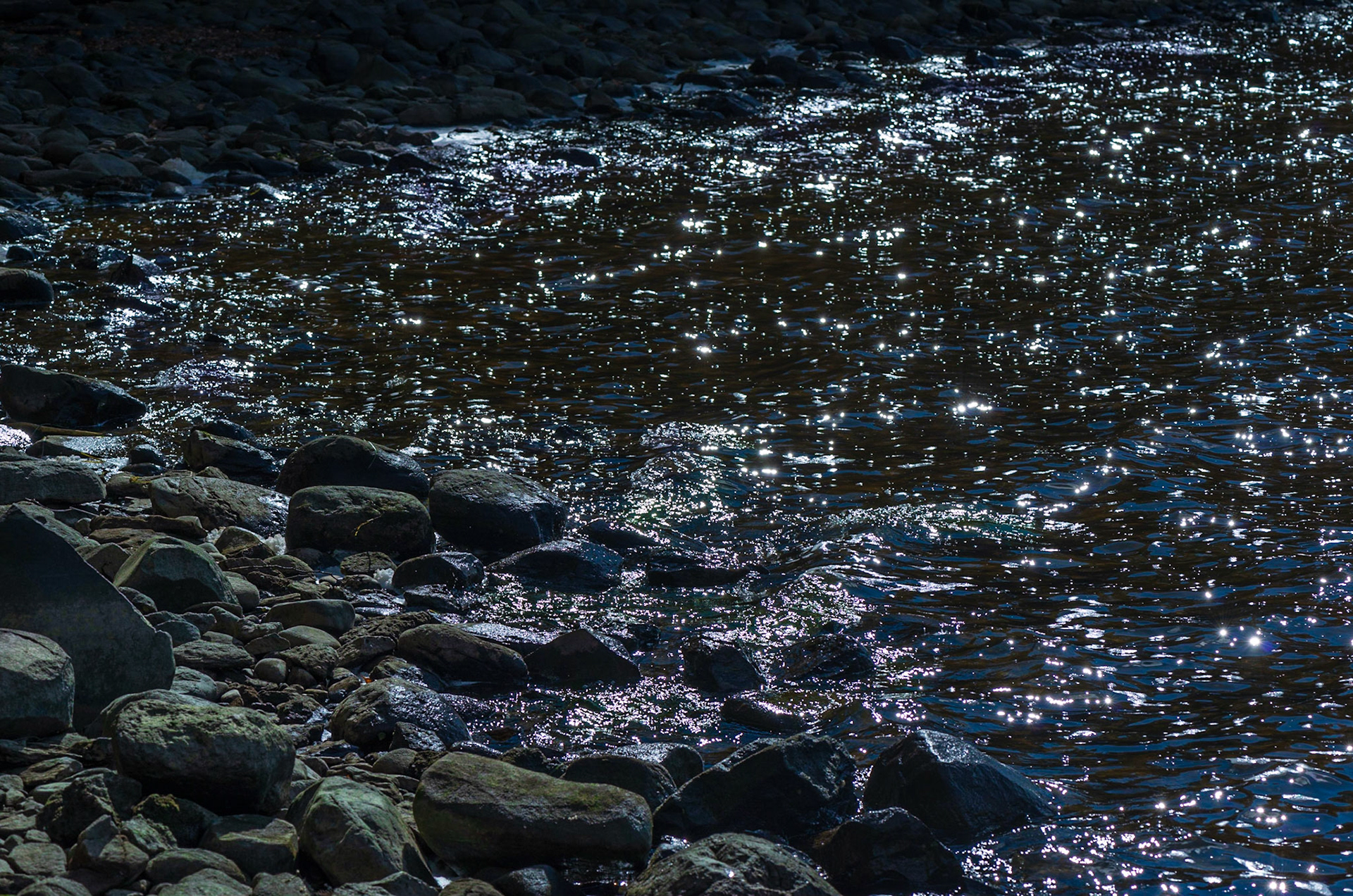 Glints of light filling the water contrast with the dark, hard rocks along the shore. Composed as a series of three triangular areas.