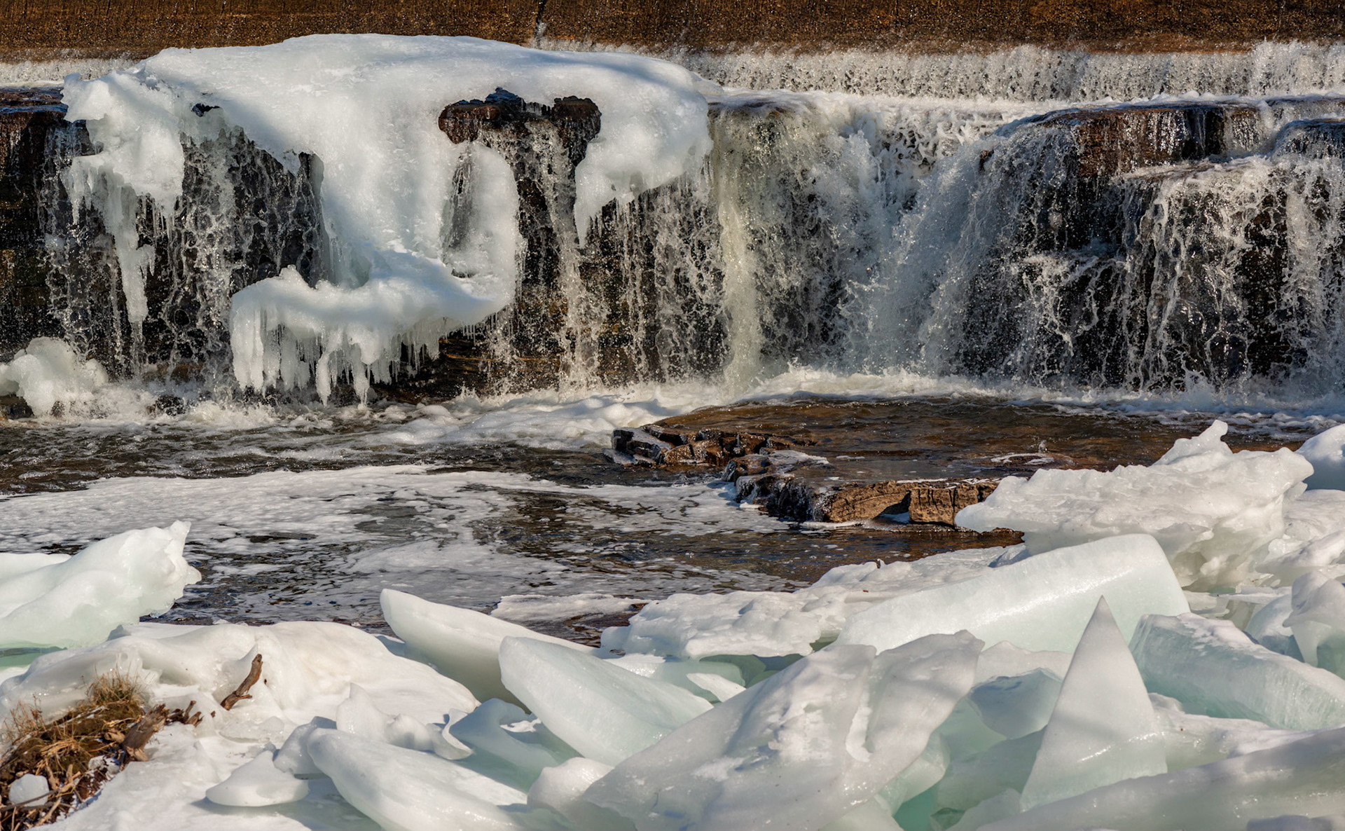 The composition consists of three horizontal layers: a foreground with interesting ice shapes, a middle ground with a flat rock for the eye to rest on, and the background of falling water.