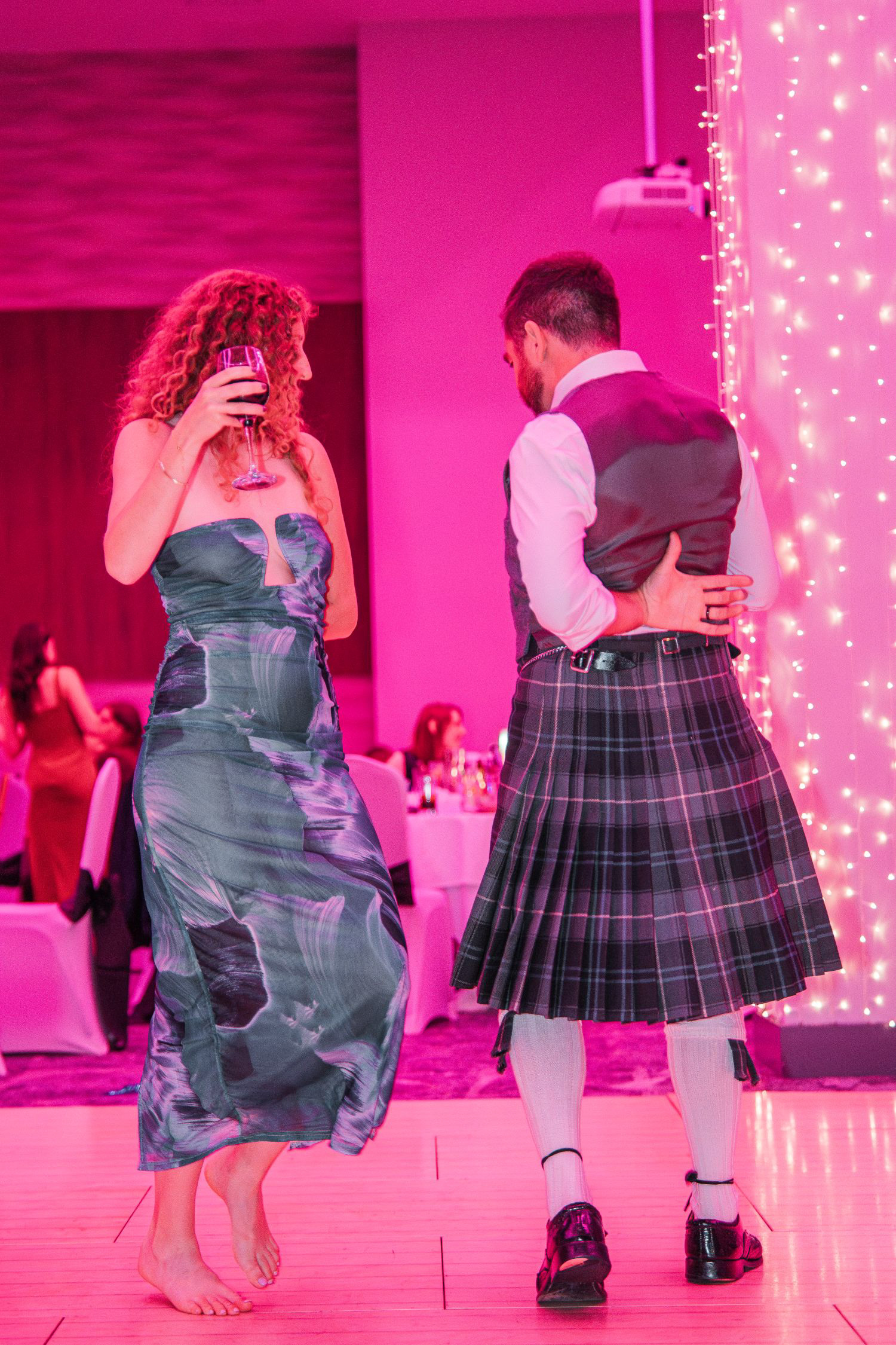 A barefoot, curly haired woman in a teal dress holds a glass of red wine as she ceilidh dances with a man in a kilt.