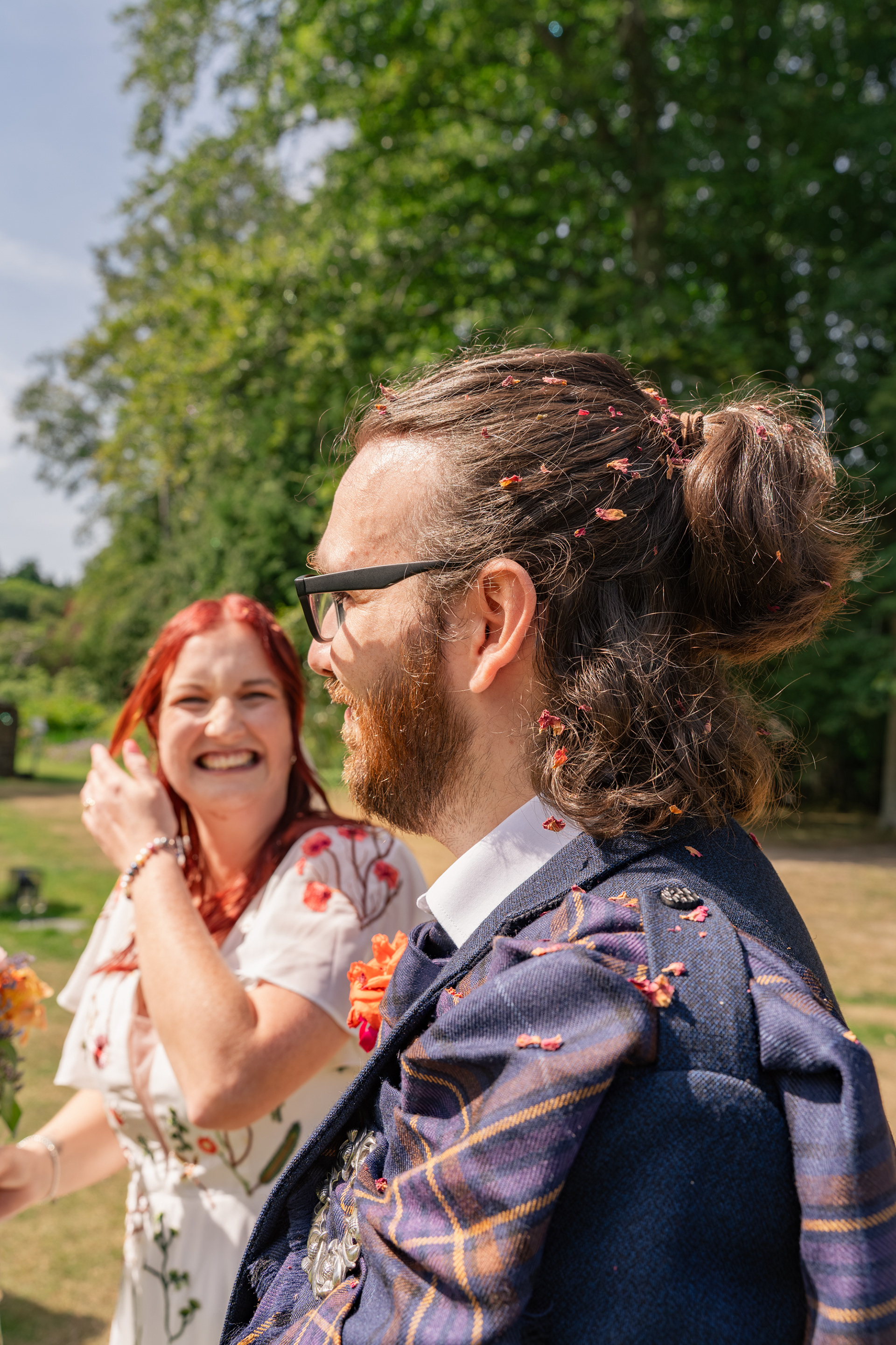 The groom's head and shoulders are covered in dried flower confetti; behind him, the bride can be seen smiling and laughing in his direction