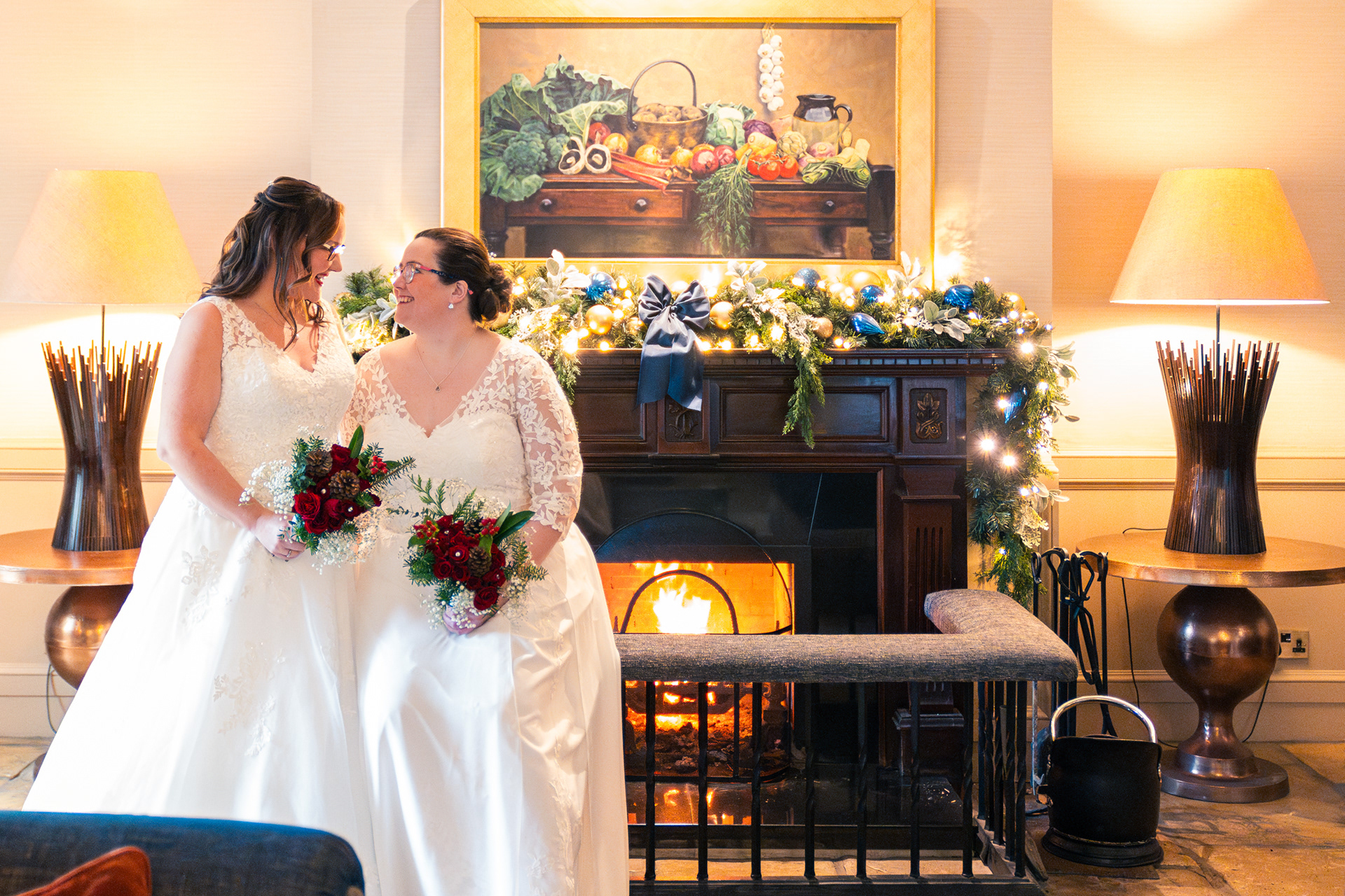 Two brides in white dresses, holding bouquets of red roses, smile at each other in front of a roaring fire, decorated for Christmas.