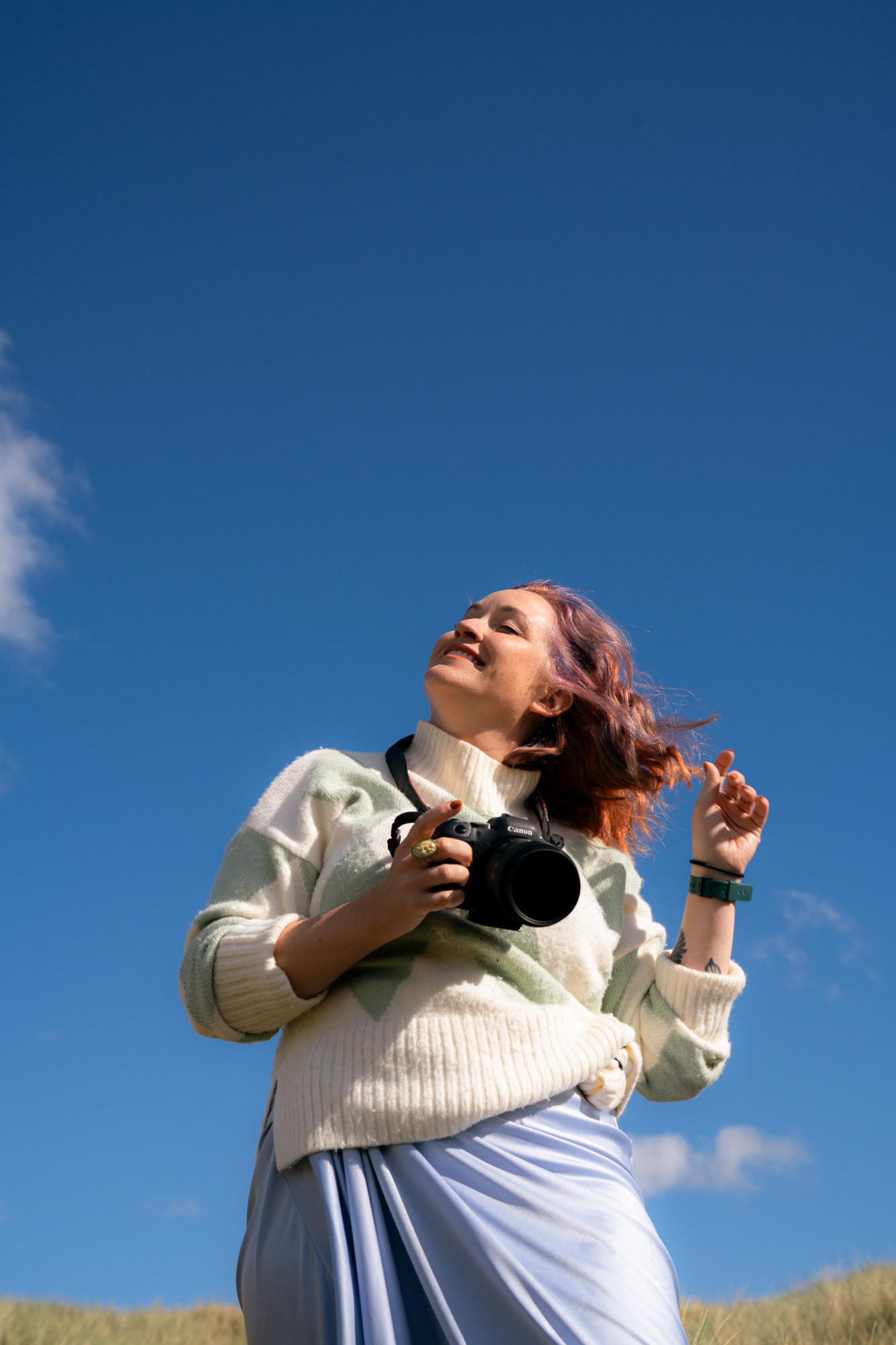 A white woman with ginger and lilac hair and freckles turns her face to the sun, eyes closed, against a bright blue sky.