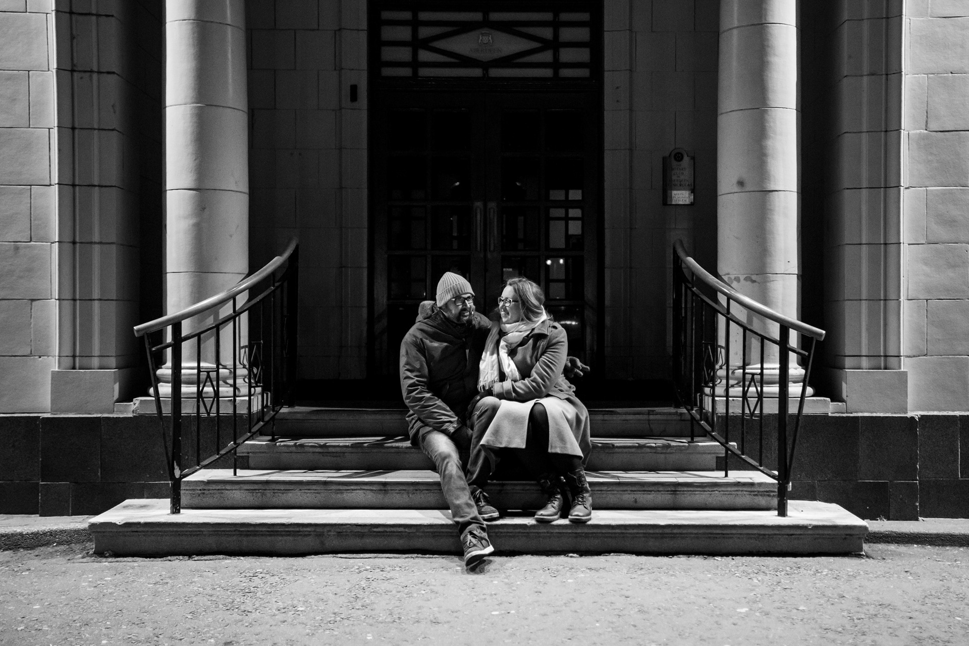 A black and white image of a cis-het couple cuddling on the steps of Aberdeen's Beach Ballroom, lit by streetlights, at 7pm on a couple's full moon photo shoot.