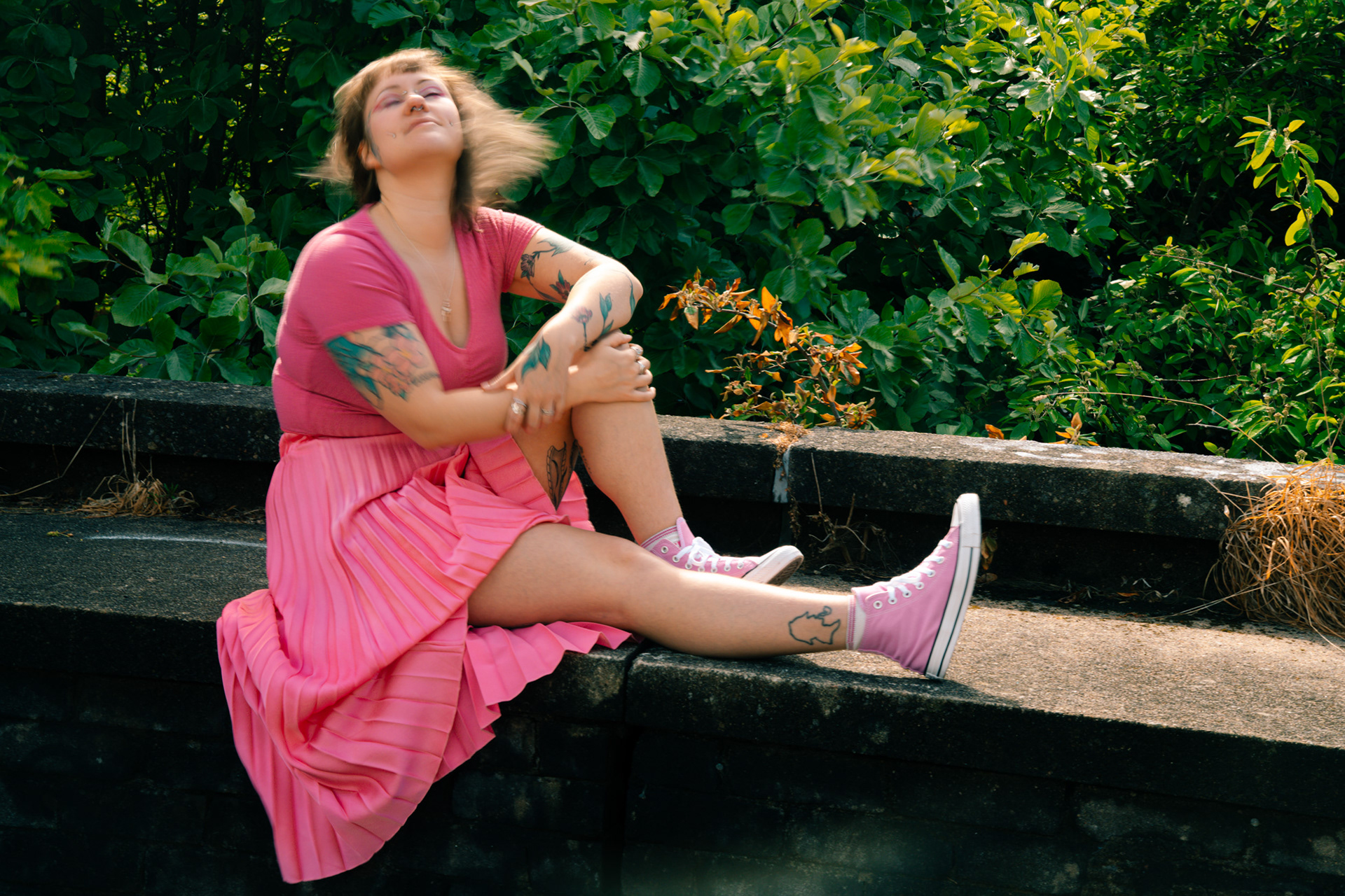 A young tattooed woman, dressed all in pink, sits on a wall in the sunshine. There are large shrubs behind her which echo the flowers up her arms. She is tossing her head, her short hair blurred by the motion.