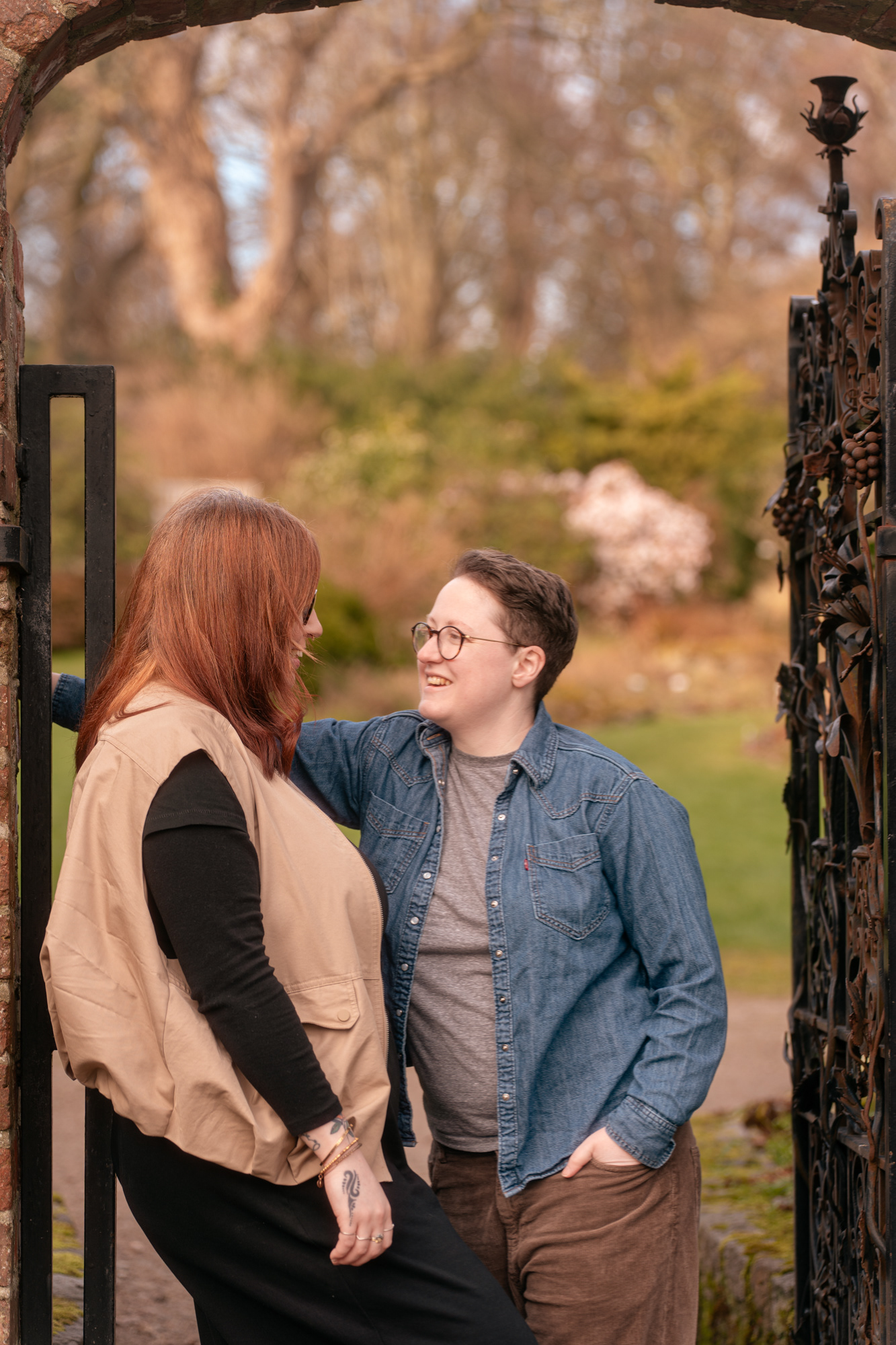 A queer couple (she/her and they/them) stand under the arch of an ornamental gate, smiling at each other, during an Aberdeen couple's photo shoot.