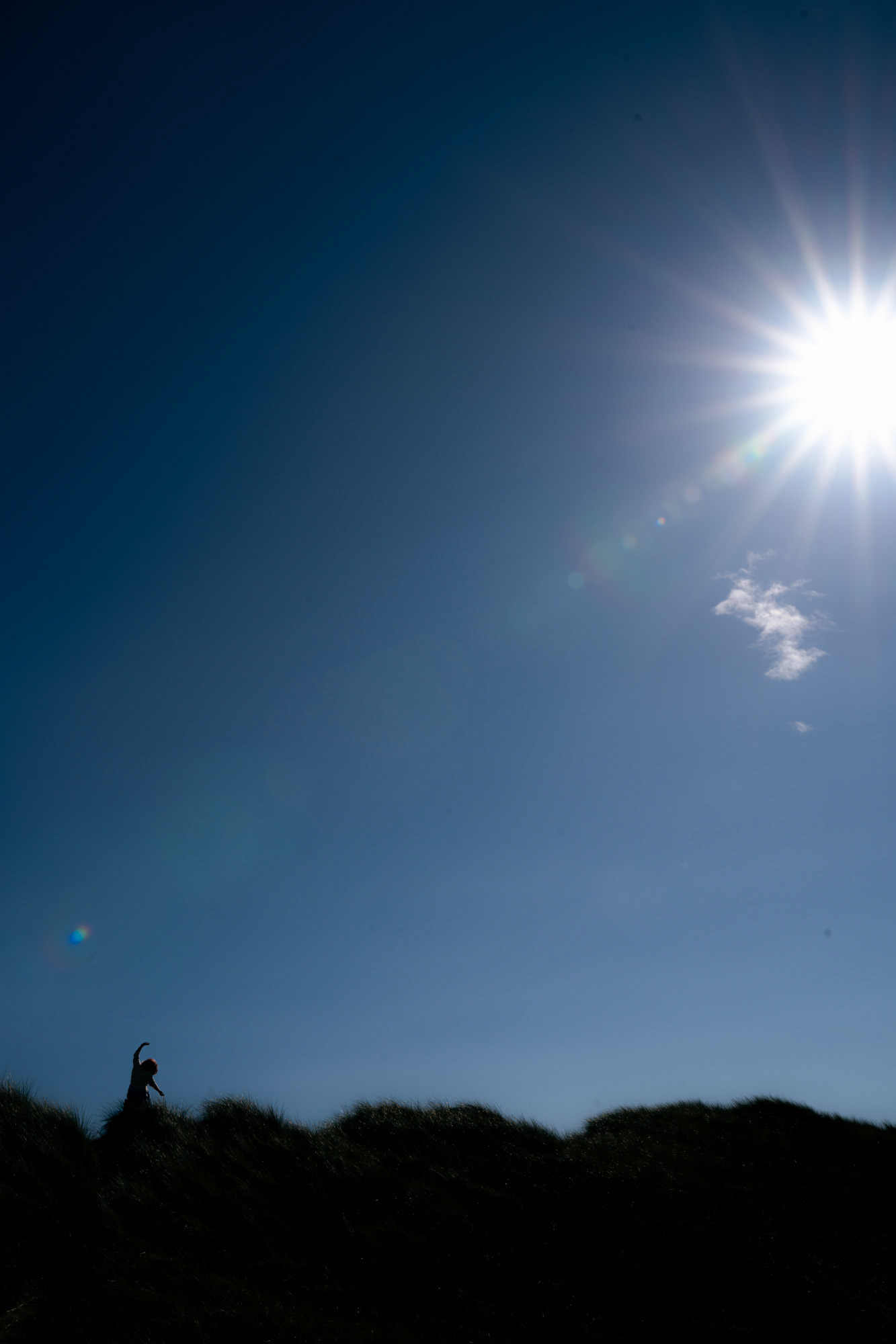 A dancing woman is silhouetted against a blue sky. She is tiny in the bottom left of the frame; the sun is large in the top right, with a sun flare streaming straight down towards her.