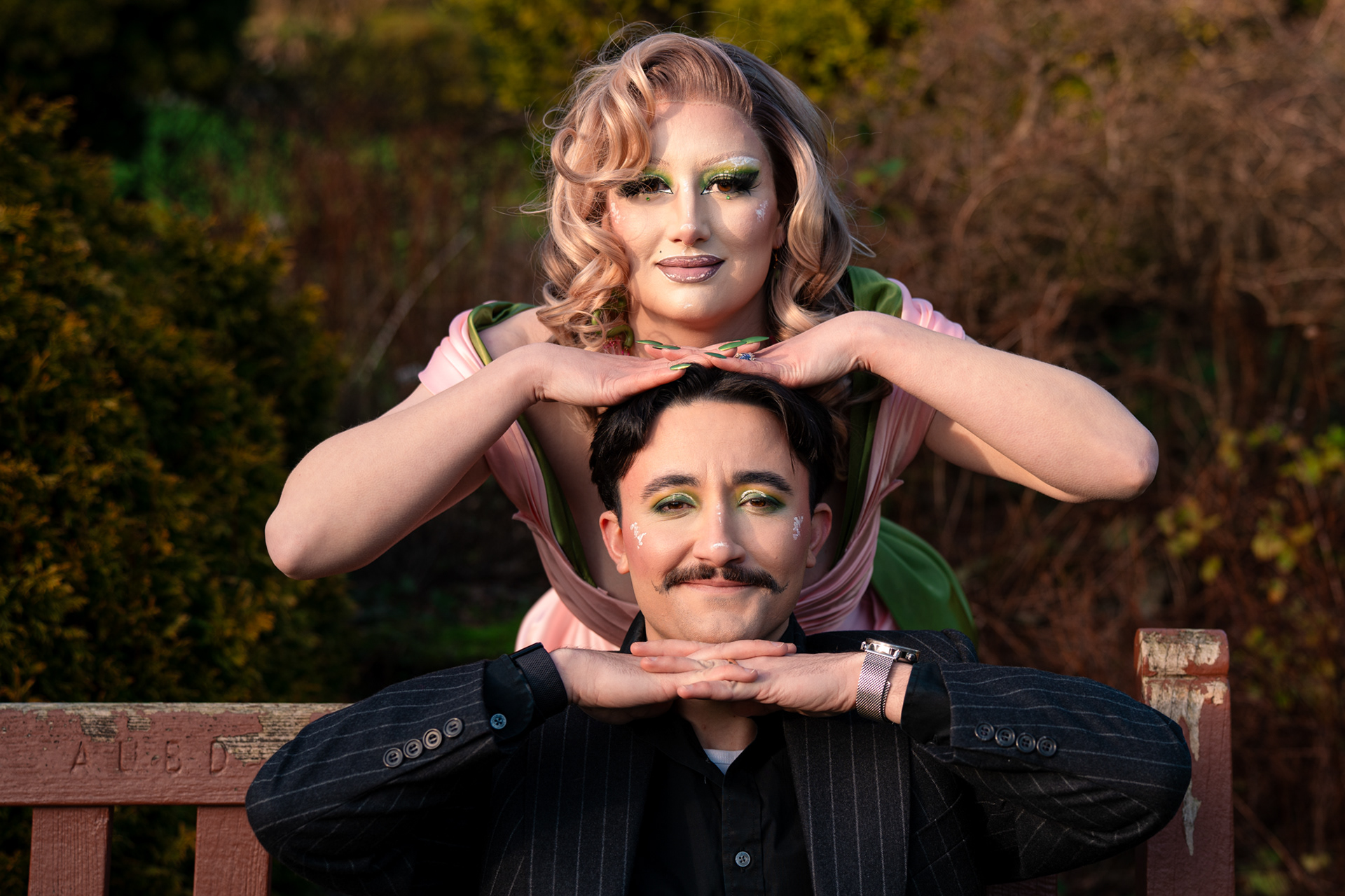 A couple in drag pose together during their Aberdeen couple's photo session. They/them is wearing a pinstripe suit and has short, dark hair and a moustache. She/her is in a glamorous pink and green dress and high green heels with dramatic makeup. Their heads are one on top of the other in an awkward pose.