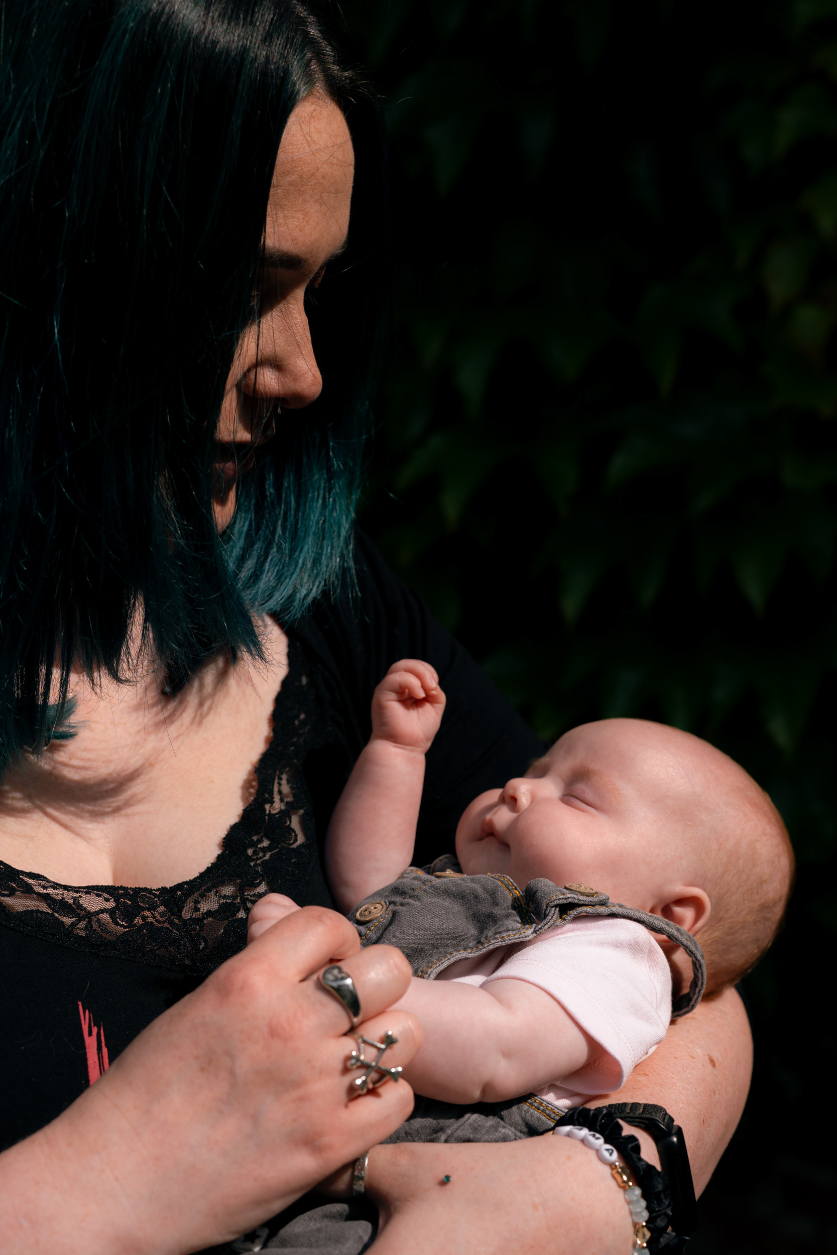 Strong contrasts in this image of a mum in black, with black and green hair, cradling and gazing down at her newborn baby. The background of ivy is thrown almost entirely into darkness.