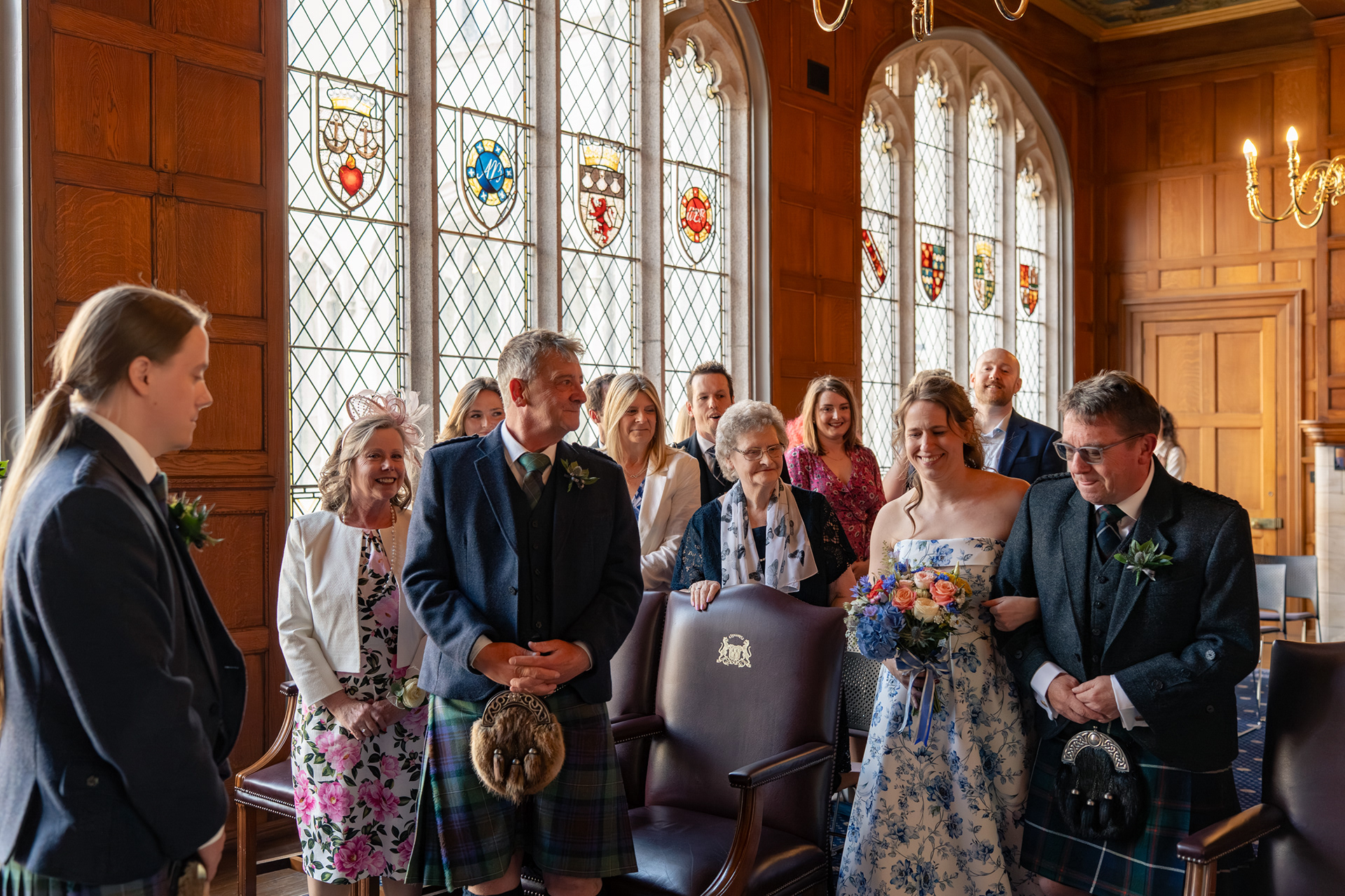 A bride in a blue floral dress is walked down the aisle of the Grant Room at Marischal College by her dad in a kilt. The long haired groom waits; guests fill the background, watching and smiling. The room is wood panelled and lit by stain glass windows and chandeliers.