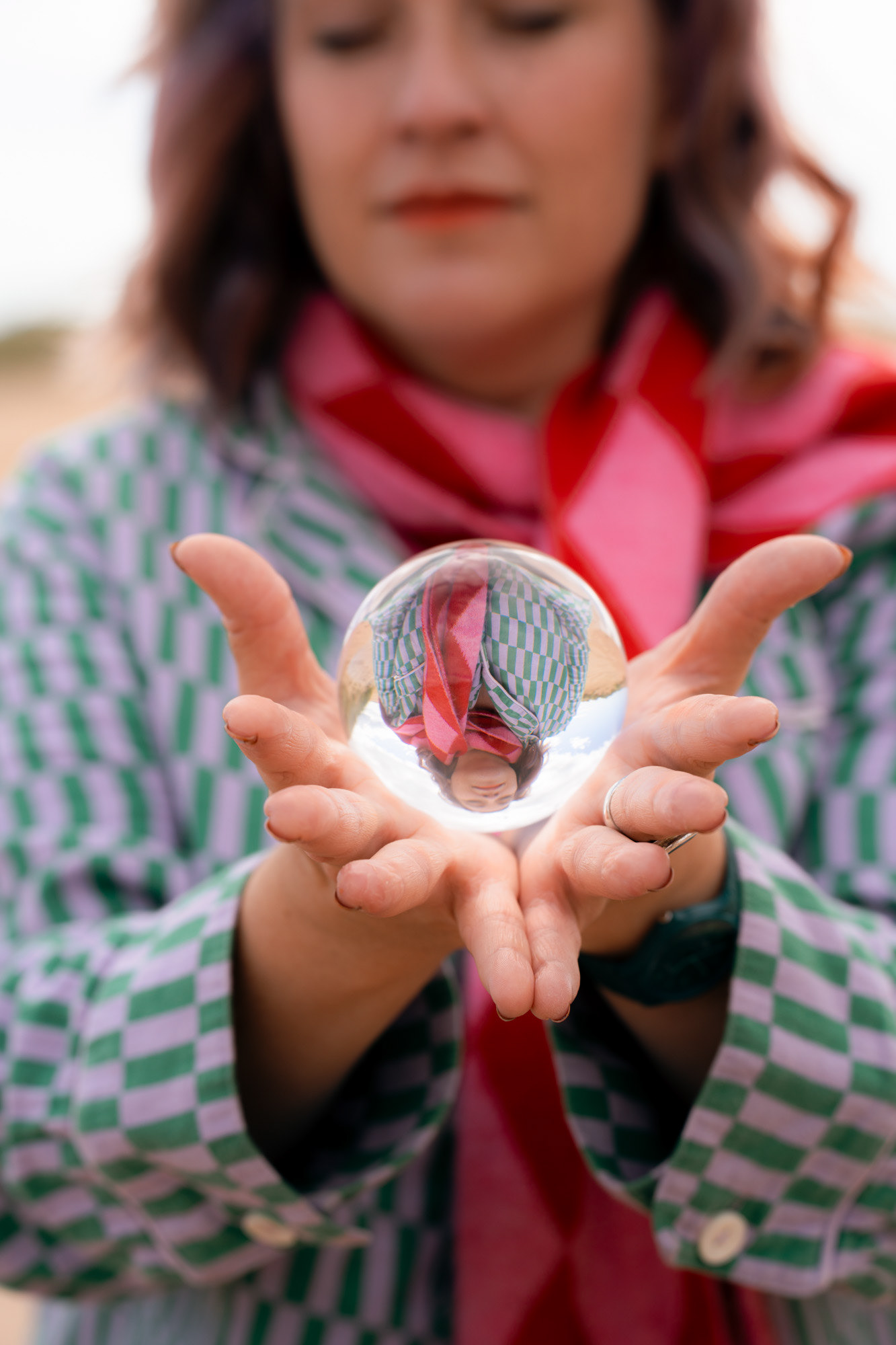 Close up of hands holding a crystal ball. A woman in a purple and green top and red and pink scarf is reflected, upside down, in the ball. The ball seems to glow against the dark backdrop of her figure.