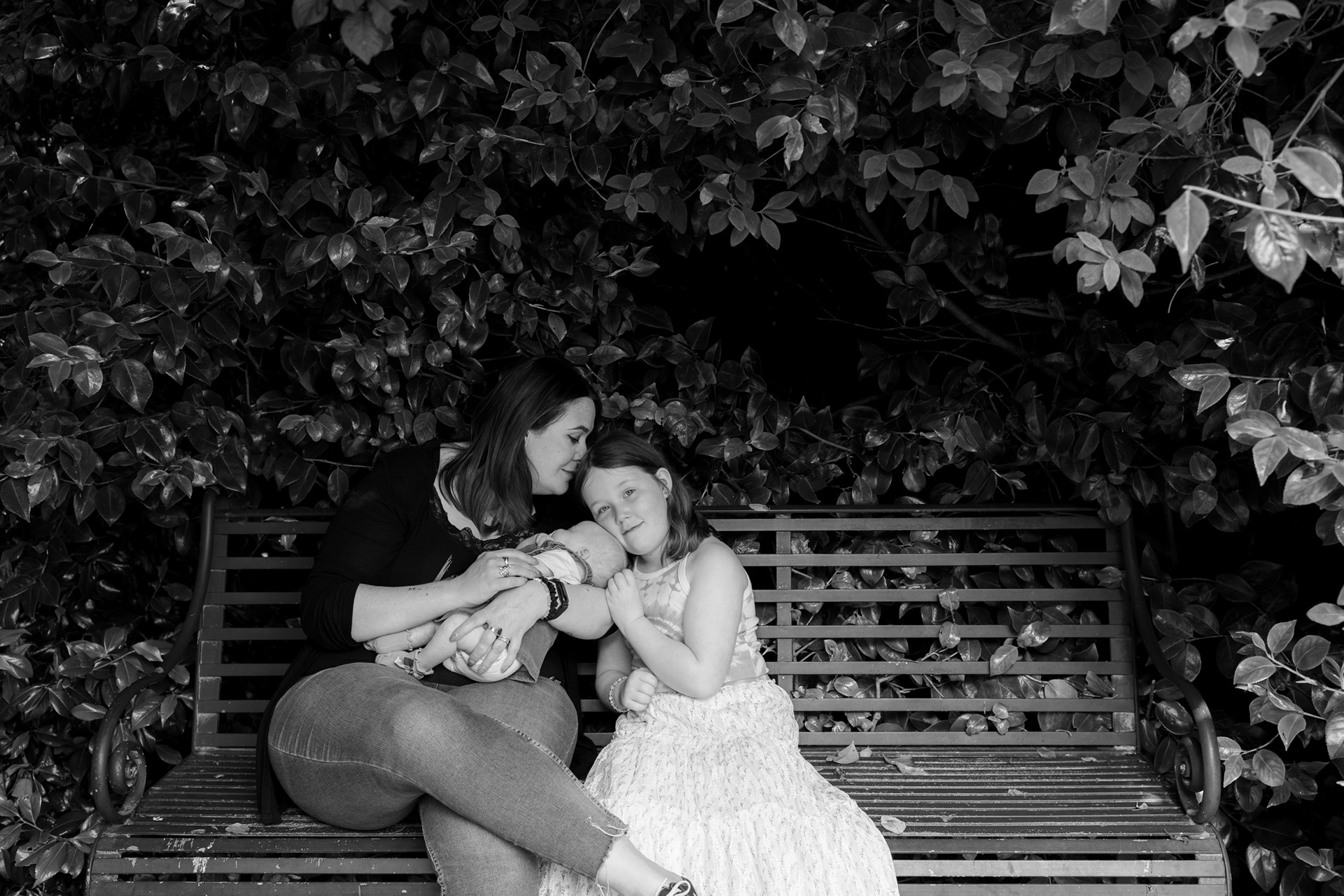 A black and white photo of mum and her two daughters - baby sleeping in her arms, 7 year old snuggling up against her. They are all on a metal park bench, completely surrounded by shrubs and climbers. Mum is leaning over to kiss her 7 year's old.